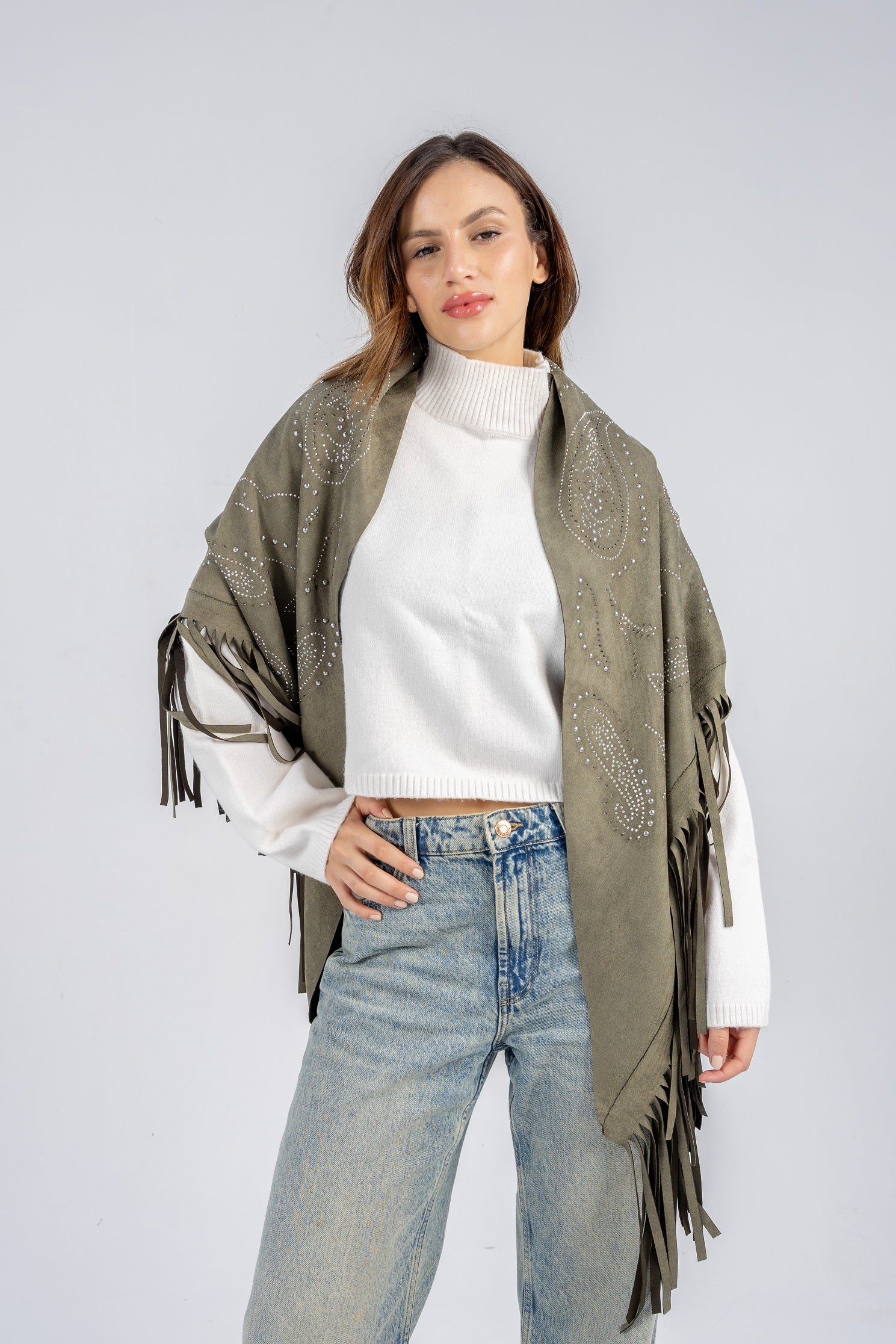 Woman wearing a green patterned shawl with fringes over a white sweater and blue jeans on a light gray background