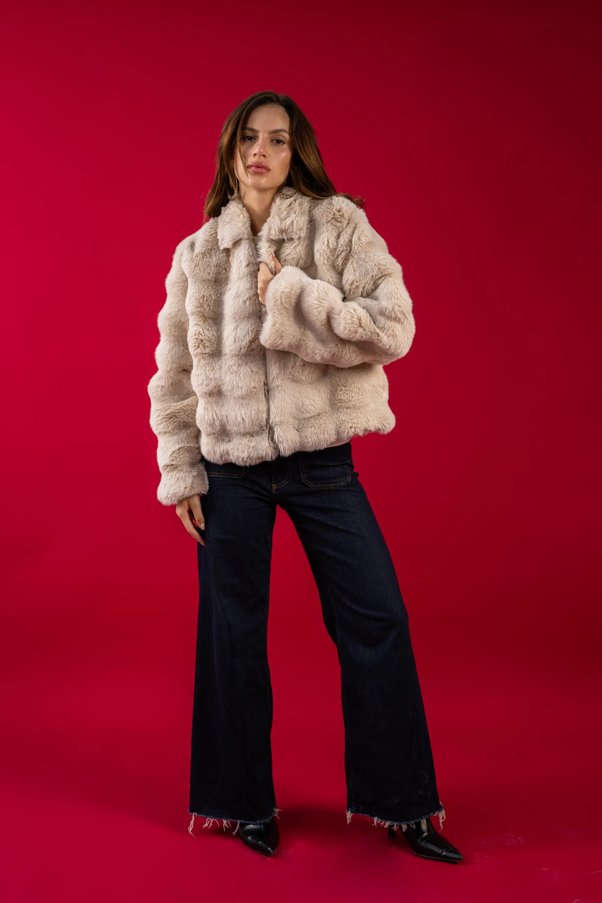 Woman wearing a beige faux fur jacket and black pants on a red background