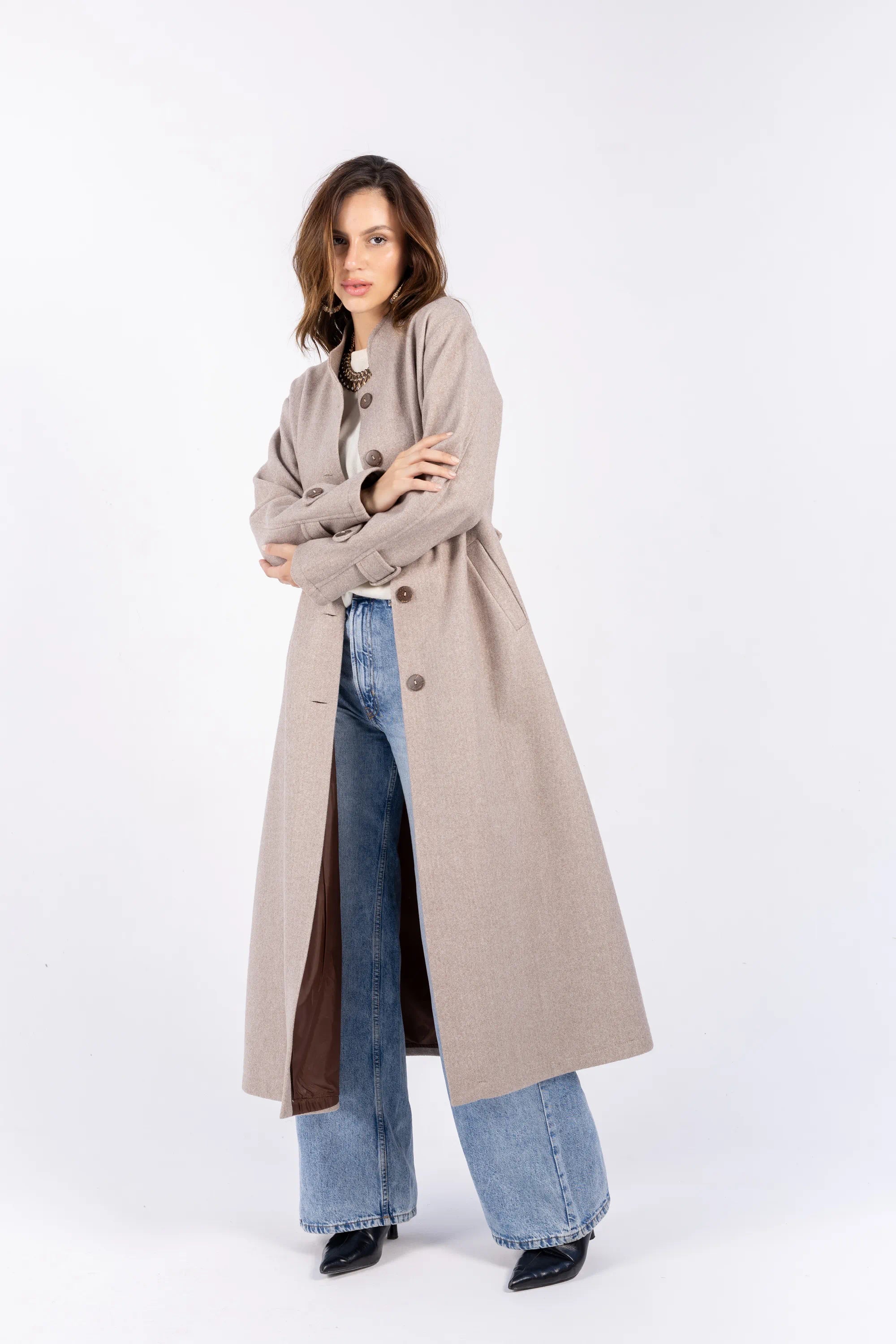 Woman wearing a long beige coat over a brown dress with blue jeans on a white background