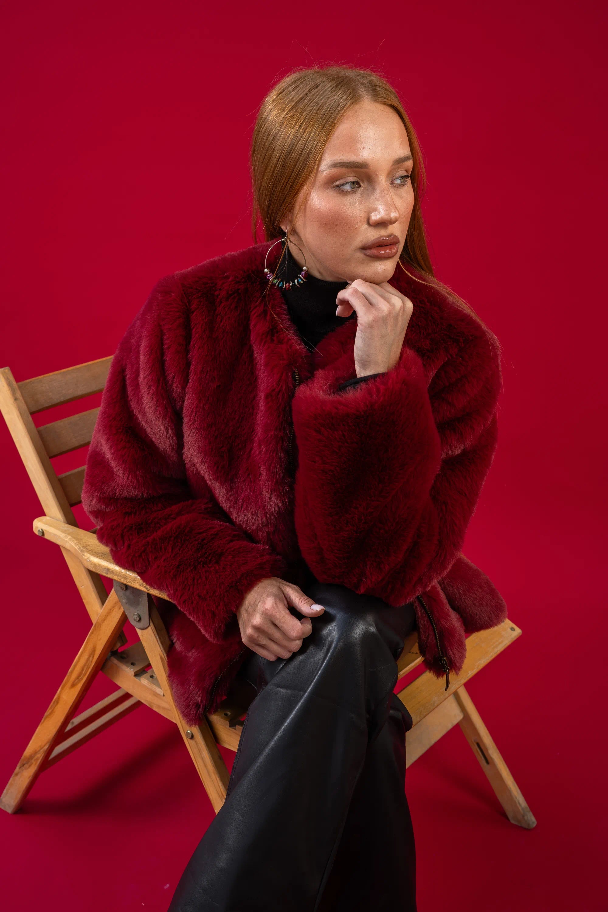 Woman in a red fur coat sitting on a wooden chair against a red background