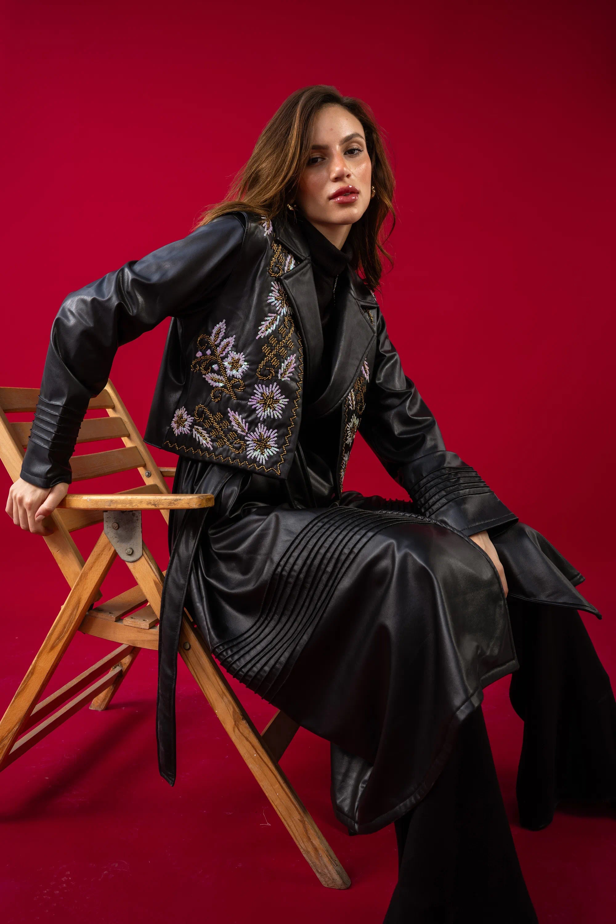 Woman in a black outfit with floral embroidery sitting on a wooden chair against a red background