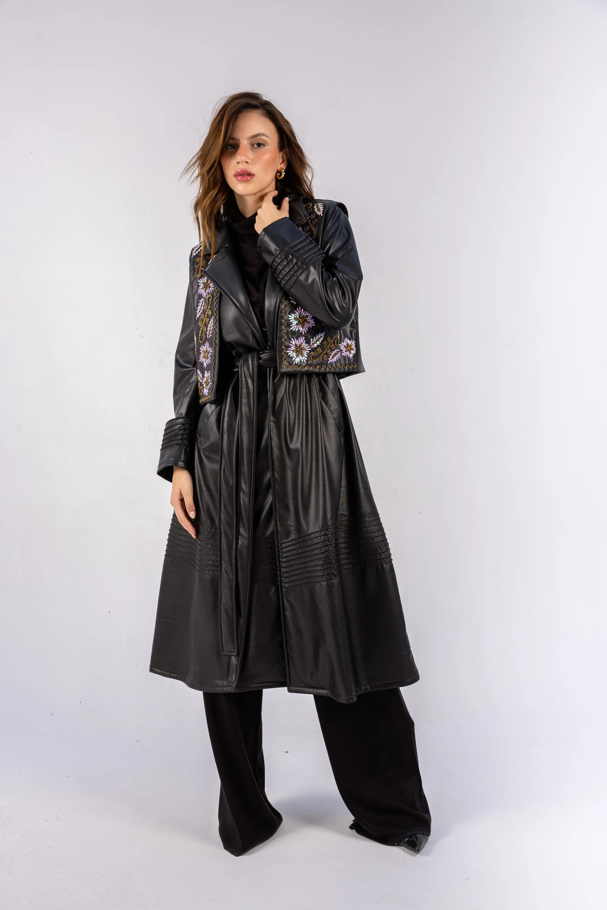 Woman wearing a long black leather coat with floral embroidery on a white background