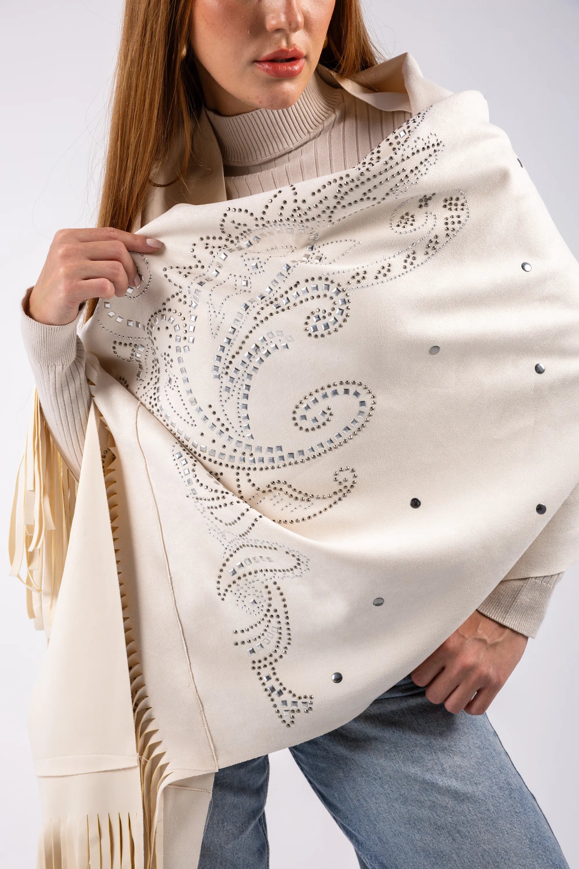 Woman wearing a cream-colored scarf with intricate patterns and fringe details on a plain background