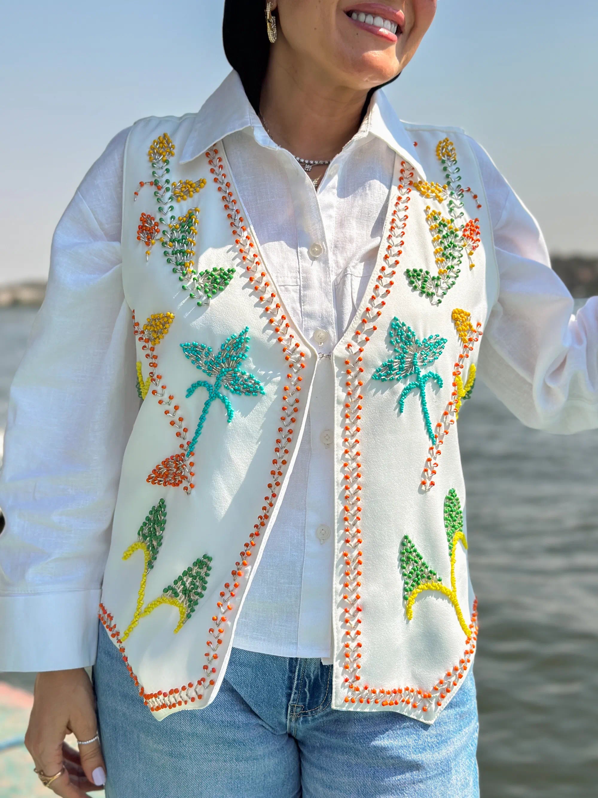 Person wearing a white embroidered vest with palm tree designs by a body of water.