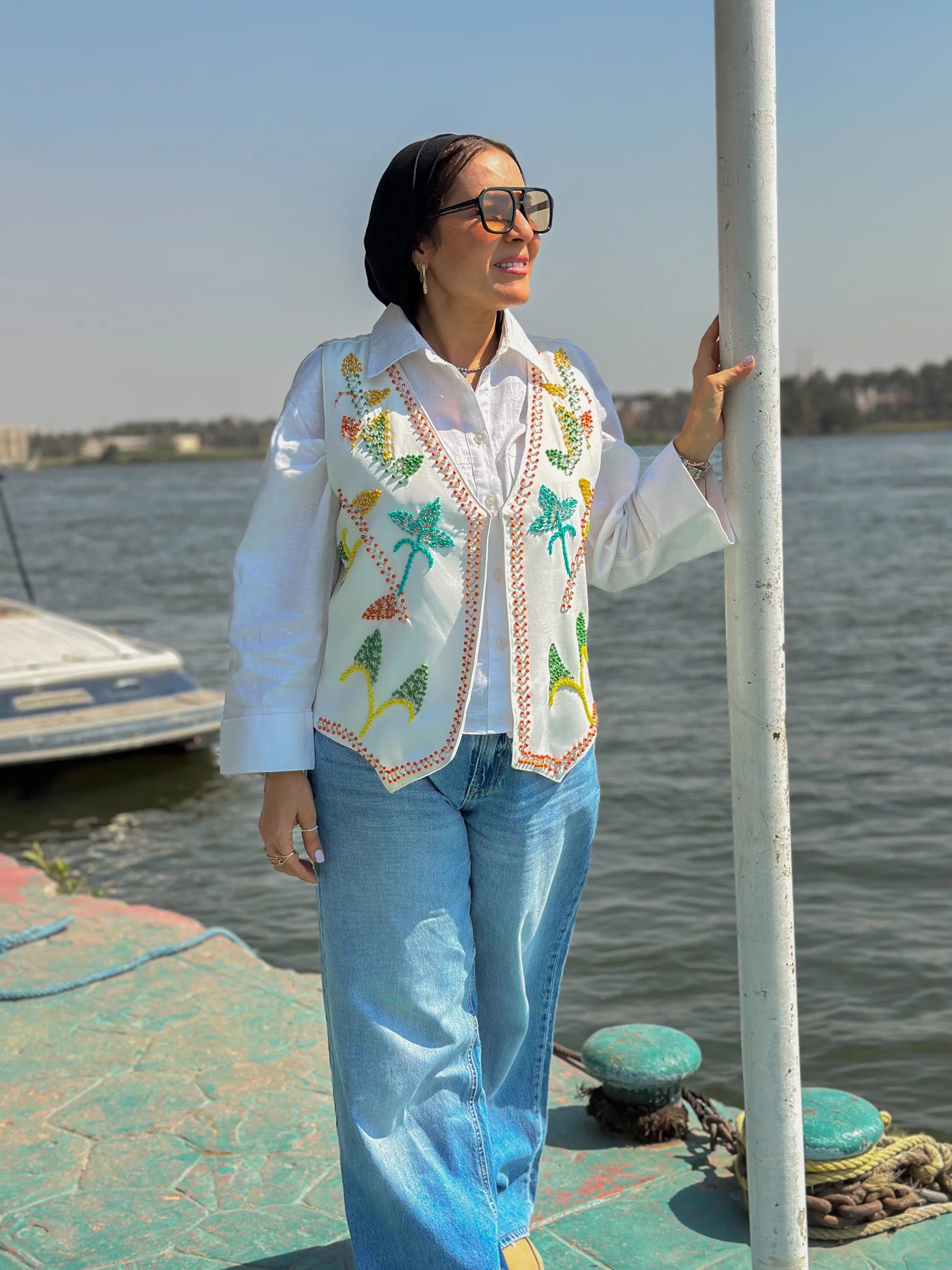 Woman standing by a body of water wearing a colorful embroidered jacket and sunglasses.