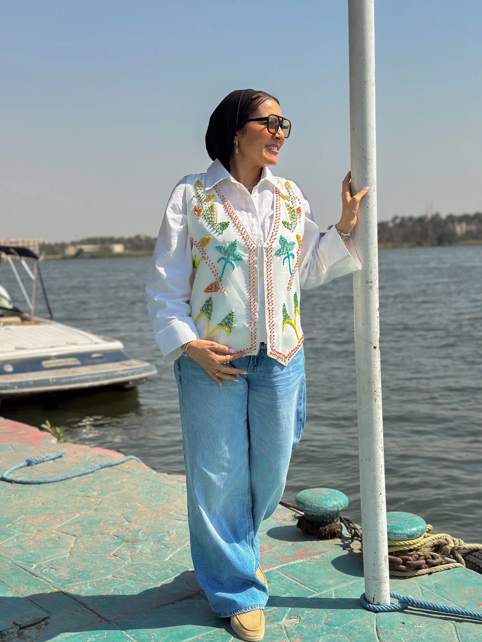 Woman standing on a dock by a body of water, wearing a colorful embroidered shirt and jeans.