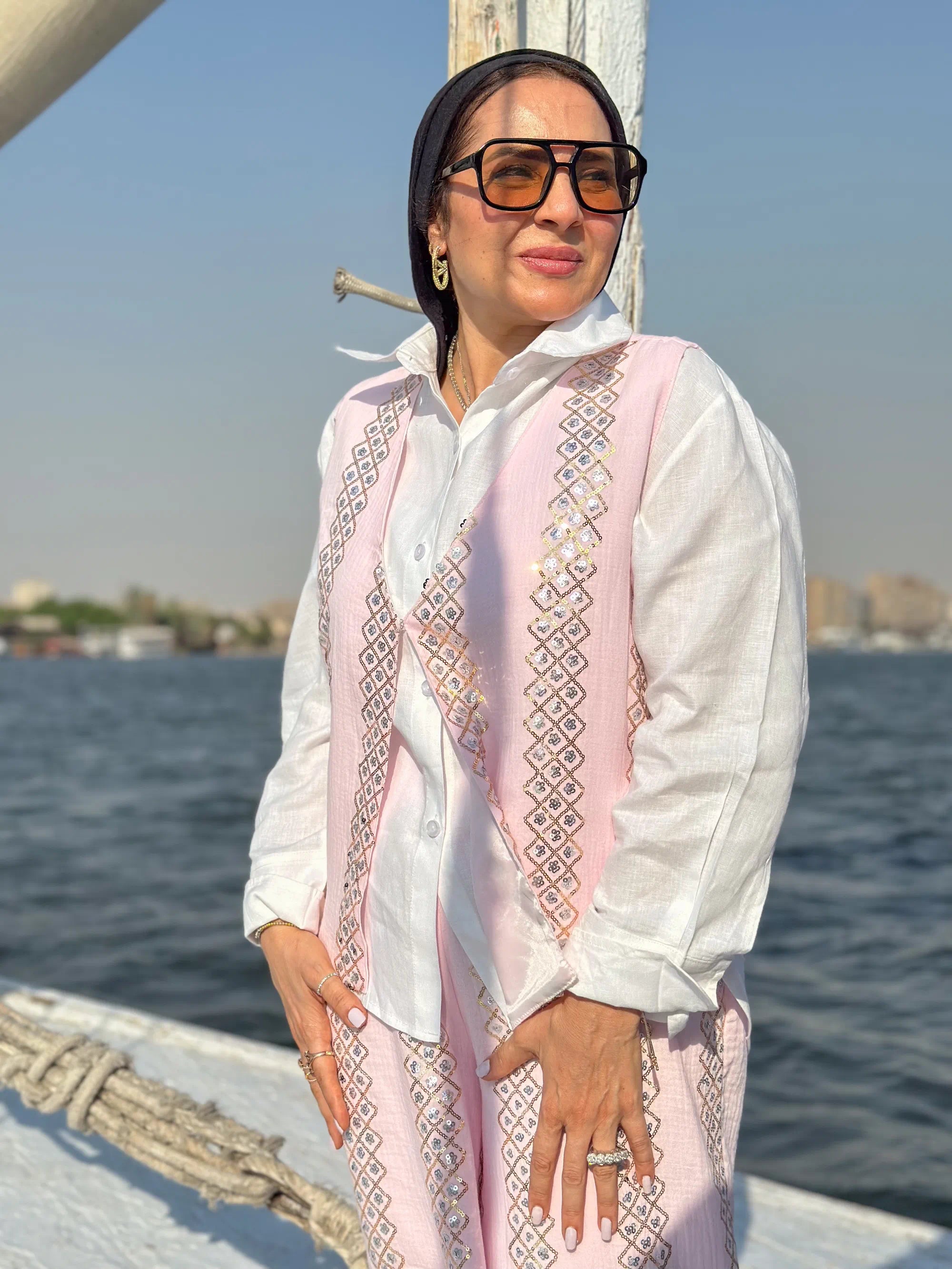 Woman in a white and pink traditional outfit standing on a boat with water and skyline in the background.