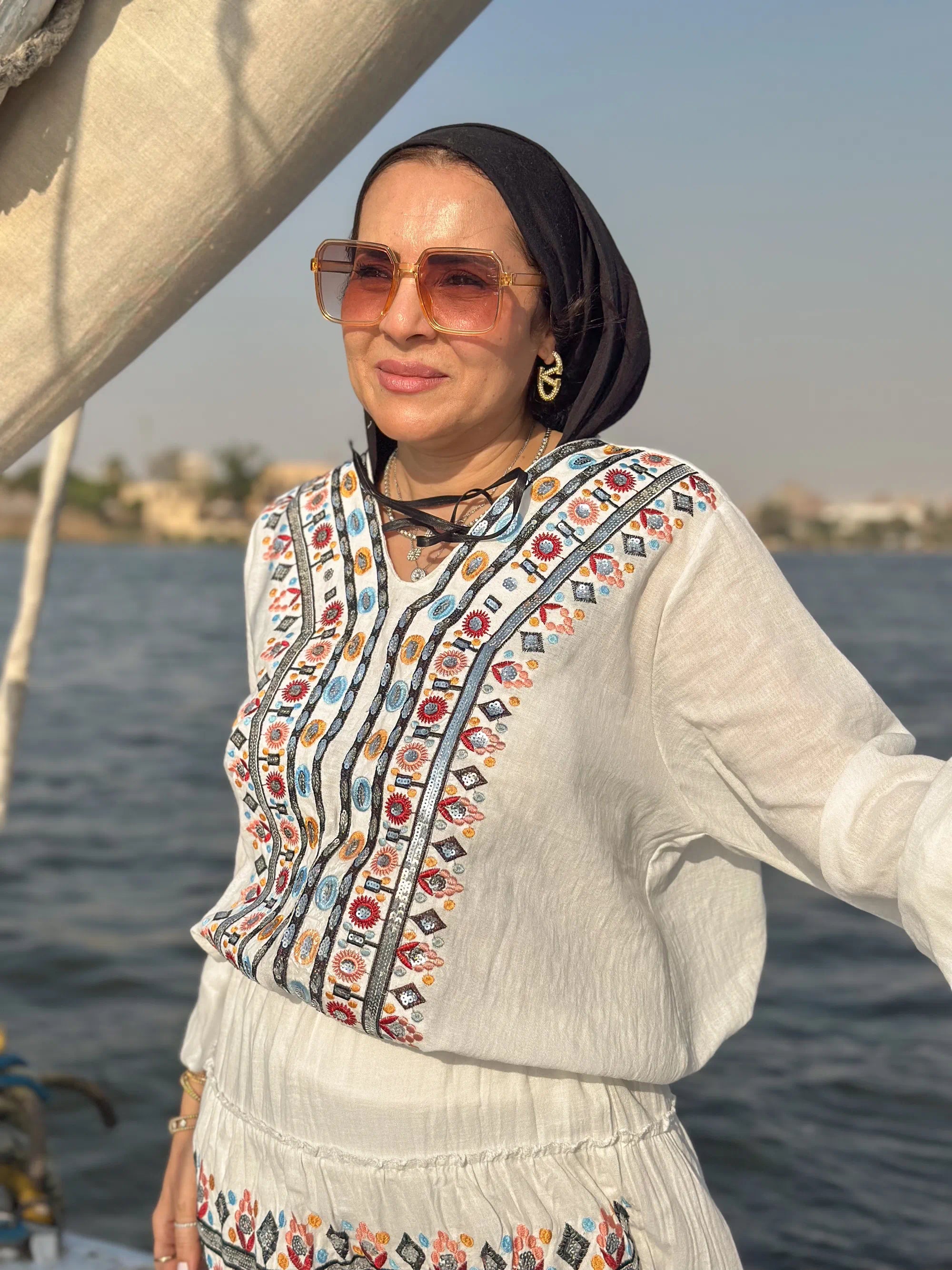 Woman wearing a colorful embroidered top and sunglasses by a body of water.
