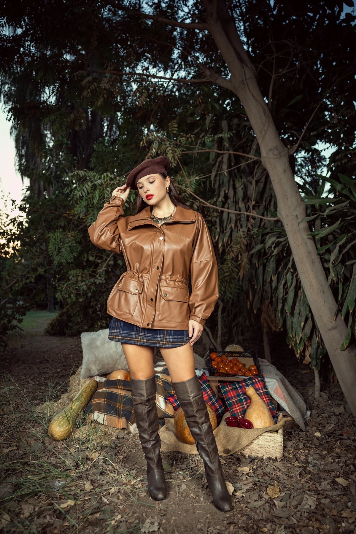 Woman in a brown leather jacket and knee-high boots standing in a forest setting.