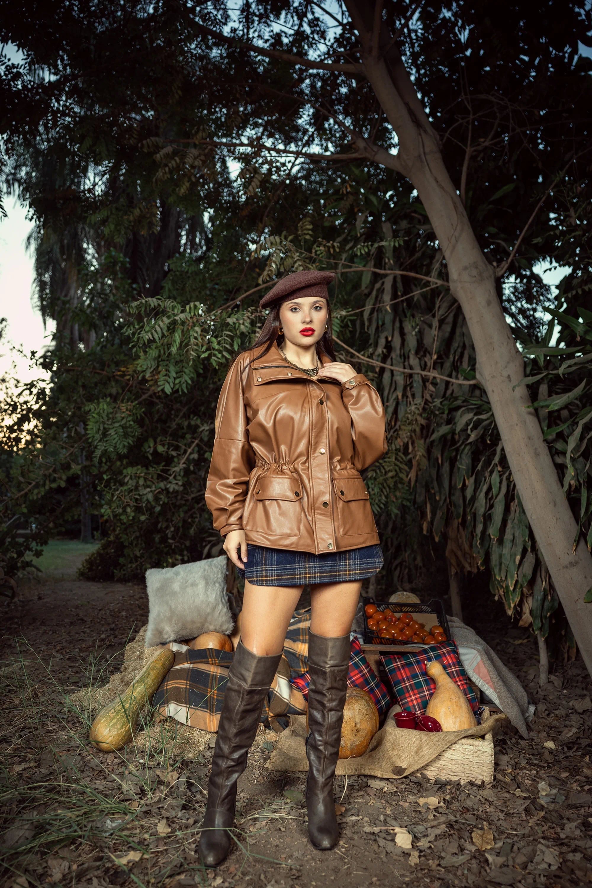 Woman in a brown leather jacket and knee-high boots standing outdoors with trees and a rustic setting.