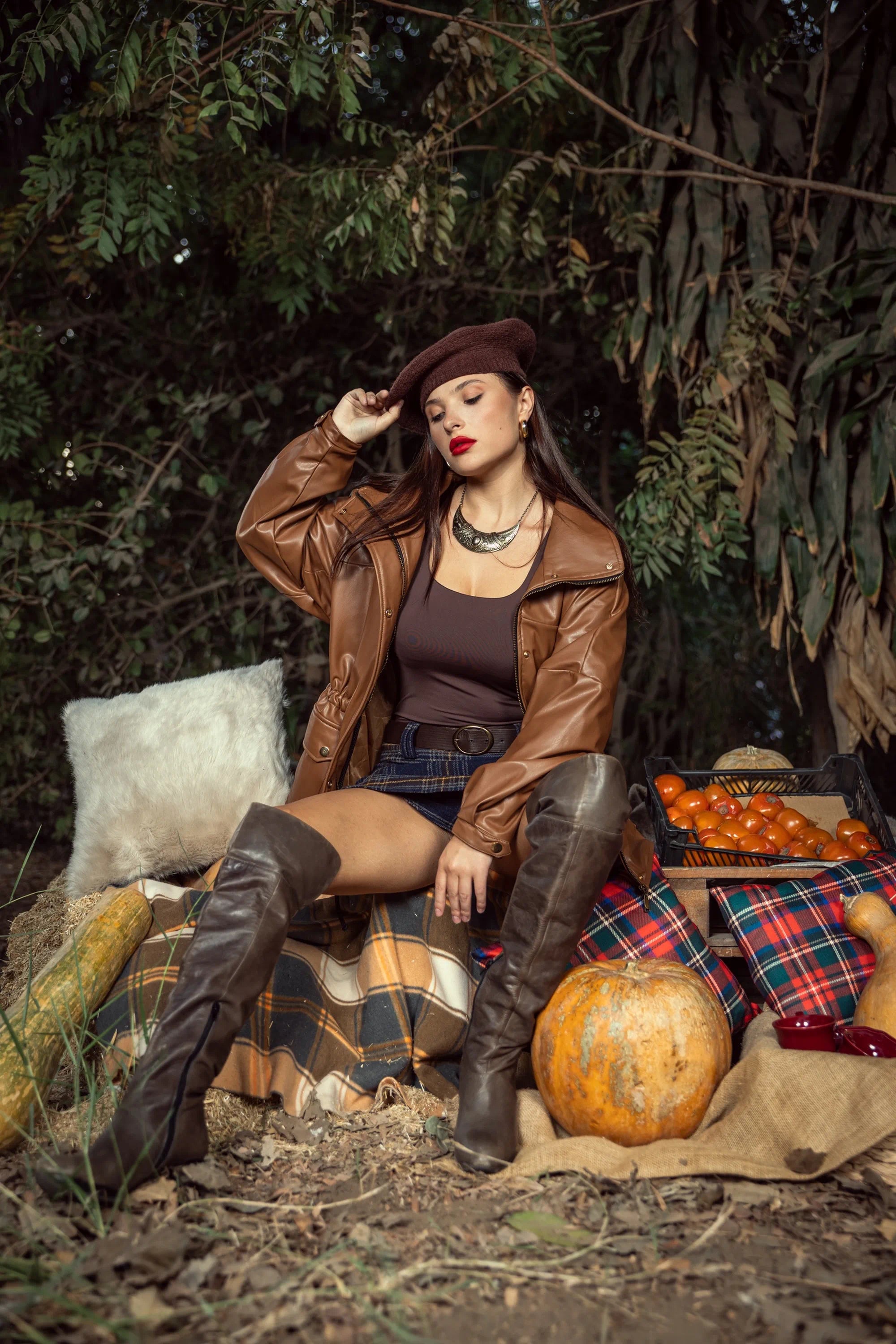 Woman in a brown leather jacket and boots sitting among pumpkins and autumn decorations.