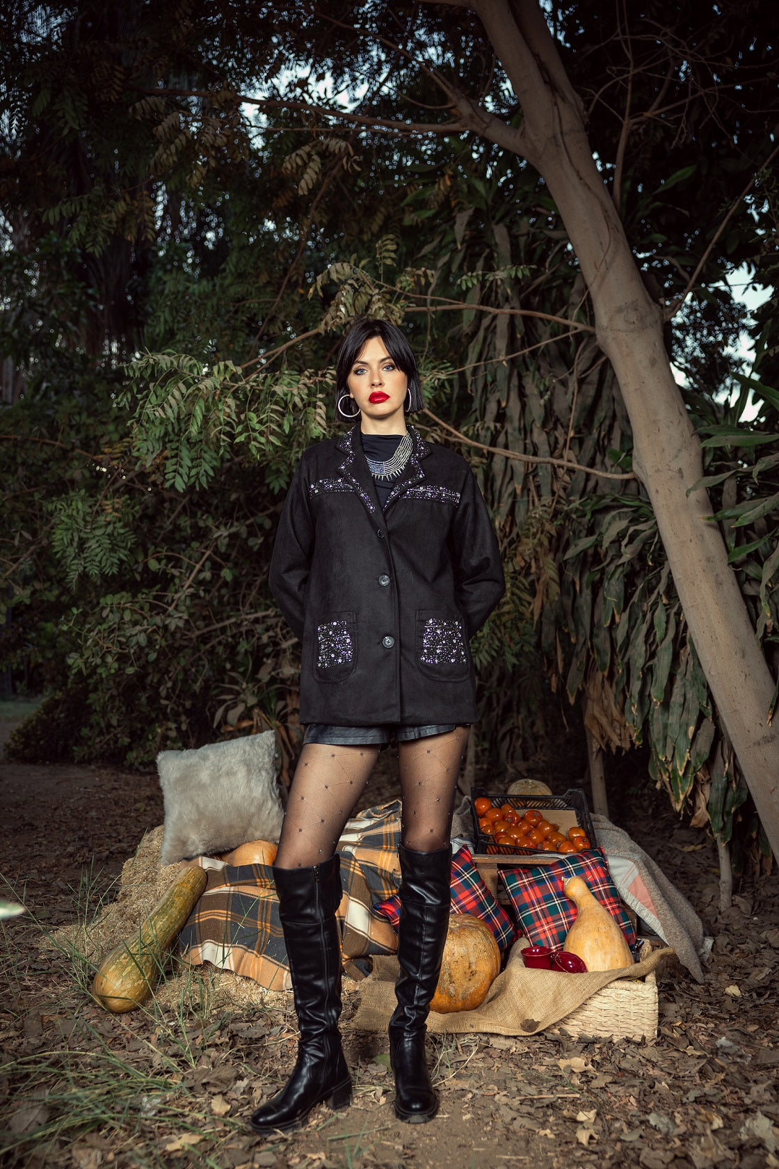 Woman in a black coat and knee-high boots standing in a forest setting with pumpkins and hay.