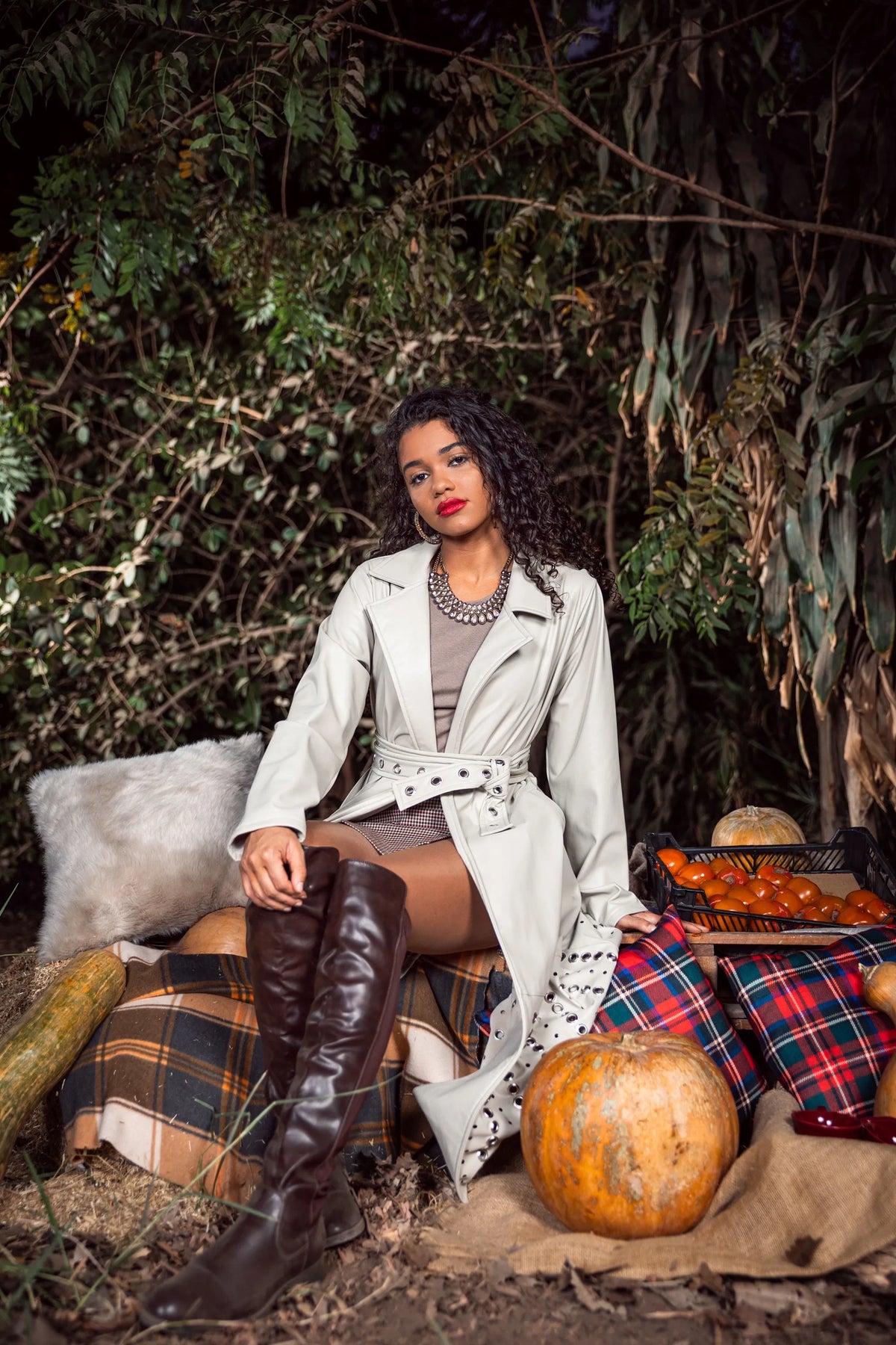 Woman in a trench coat and knee-high boots sitting outdoors with pumpkins and plaid blanket.