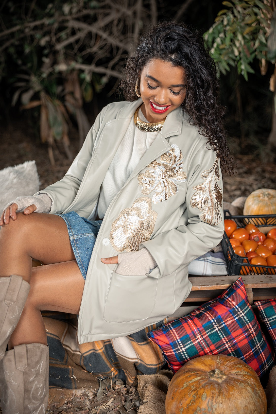Woman sitting outdoors with pumpkins and a basket of oranges, wearing a light-colored coat with sequin details.