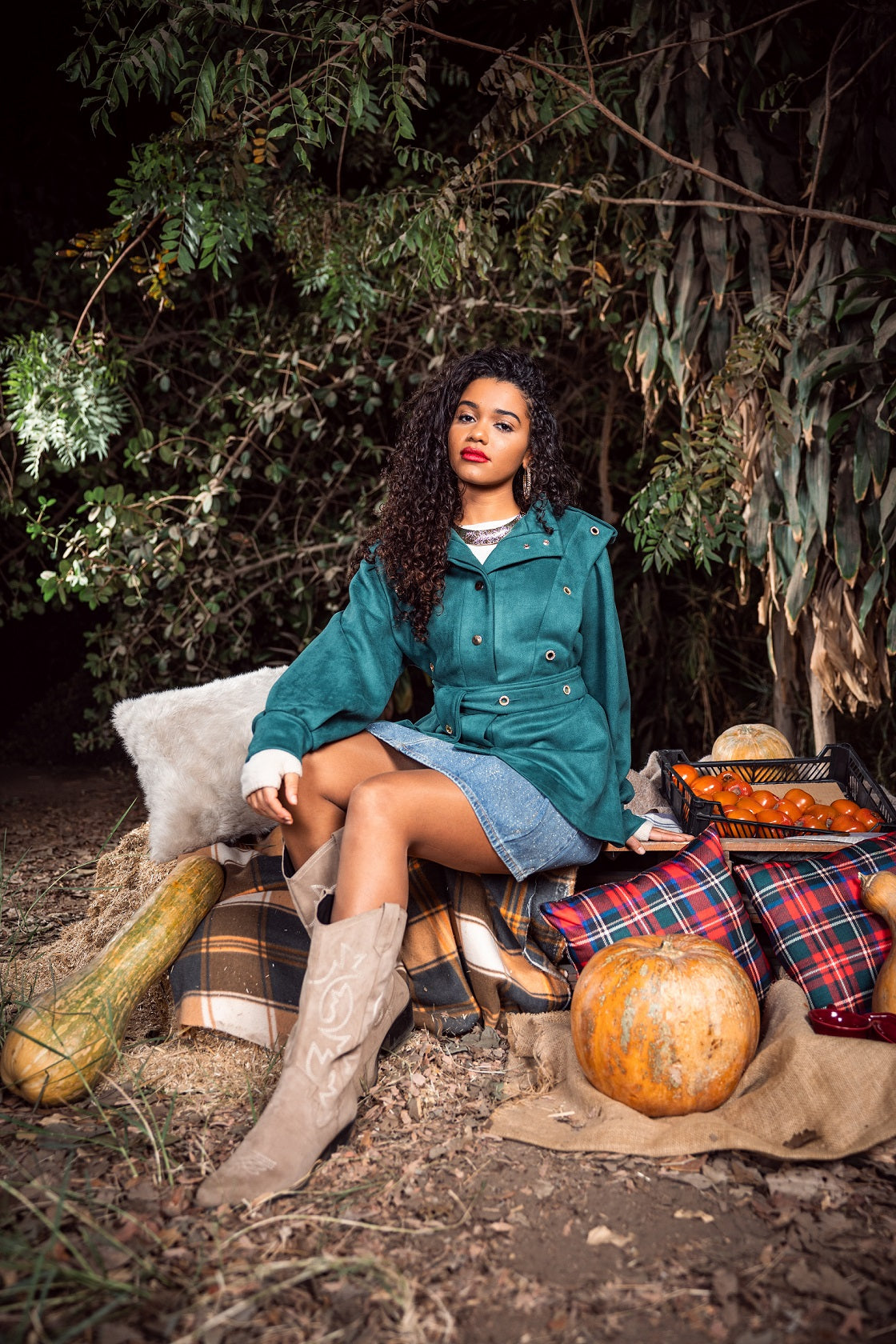 Woman in a teal coat sitting outdoors with pumpkins and a plaid blanket