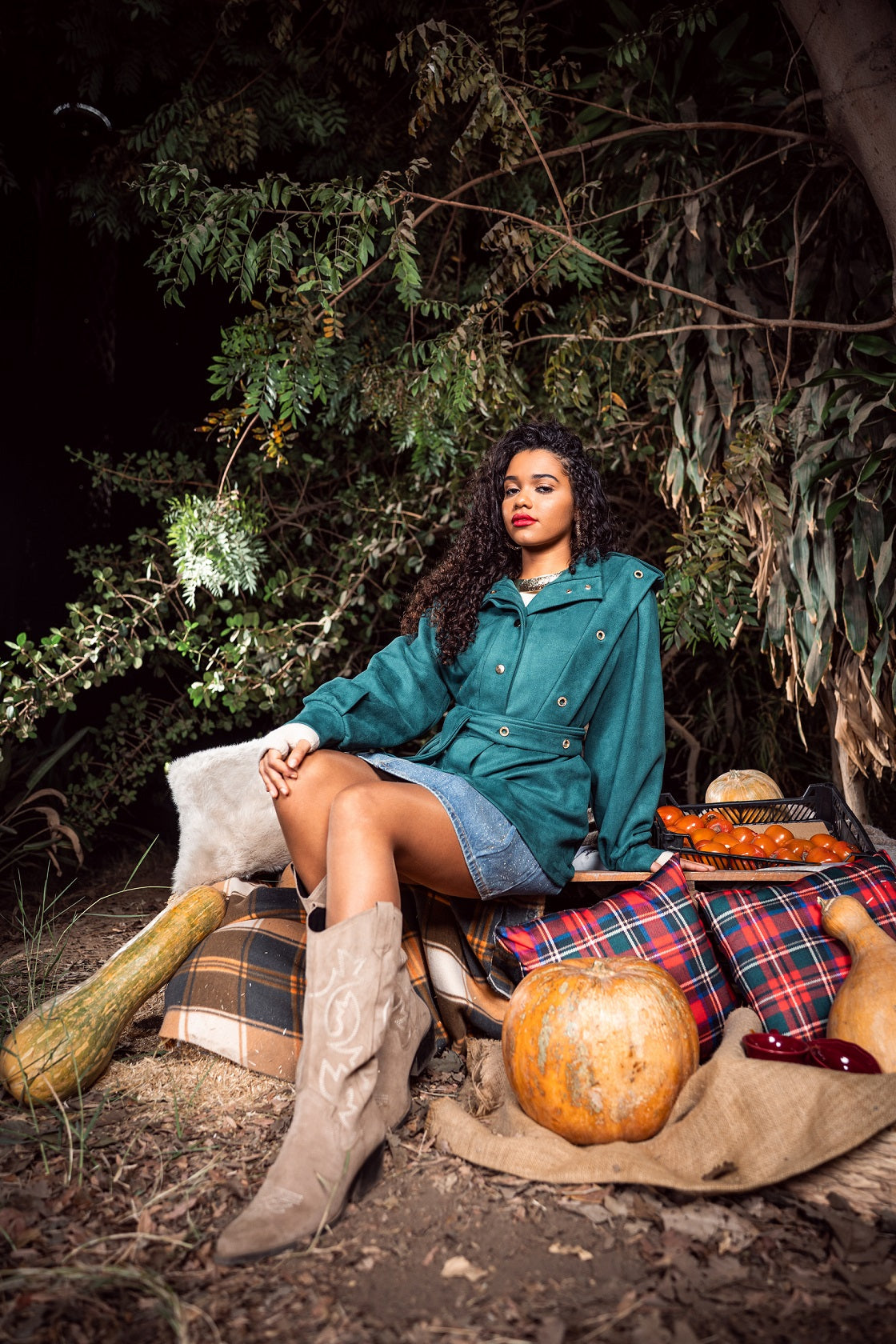 Woman in teal jacket and plaid skirt sitting outdoors with pumpkins and hay.