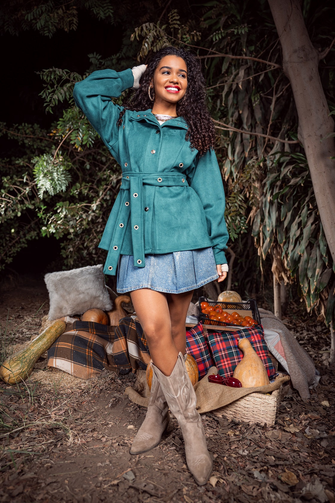 Woman in teal coat and denim skirt standing in a forest setting with plaid blanket and bag.