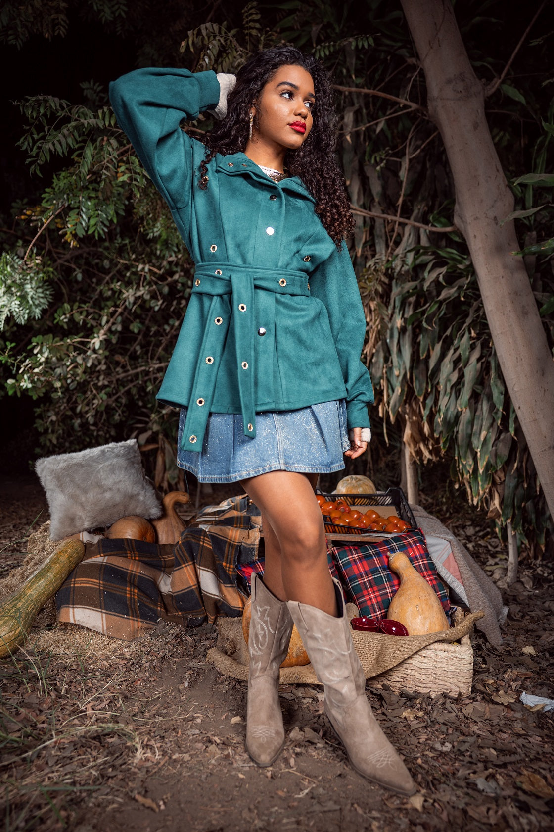 Woman in teal coat and denim skirt standing in a forest setting with rustic elements.