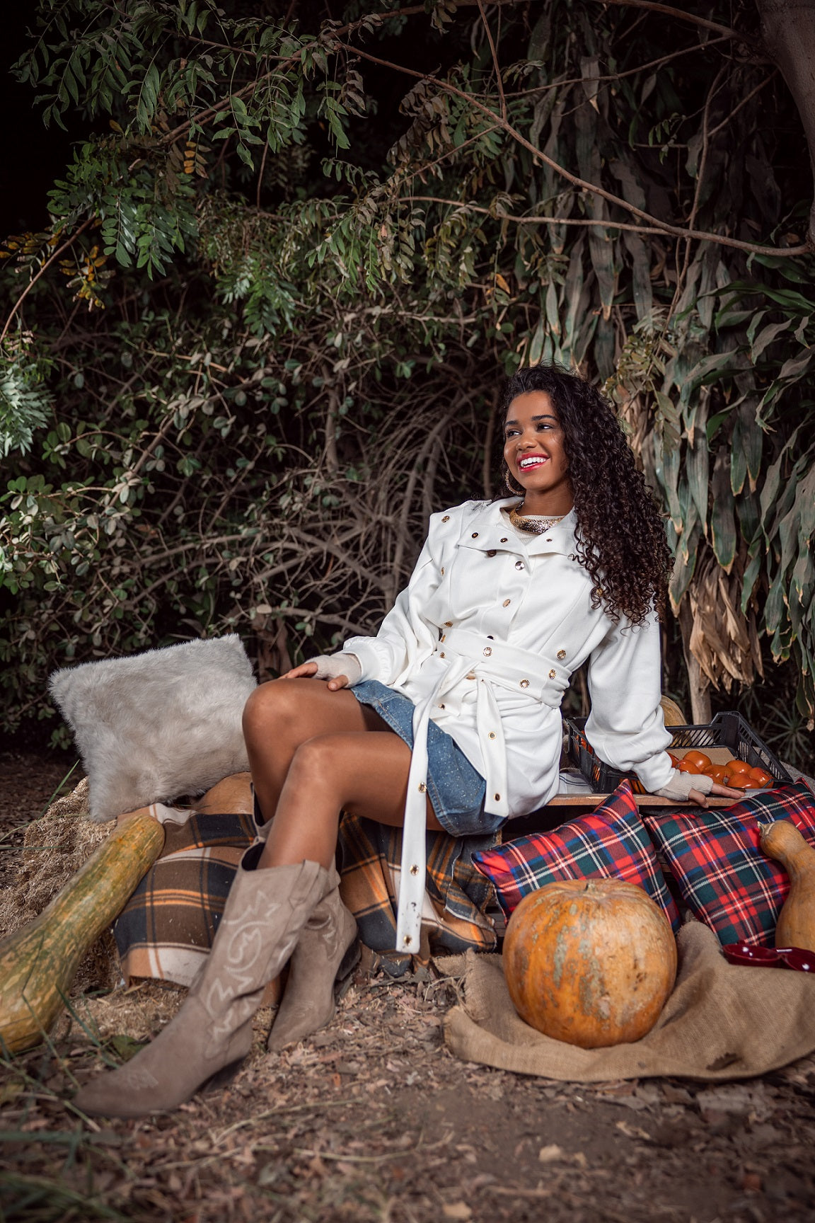Woman sitting outdoors with pumpkins and plaid blanket, wearing a white jacket and brown boots.