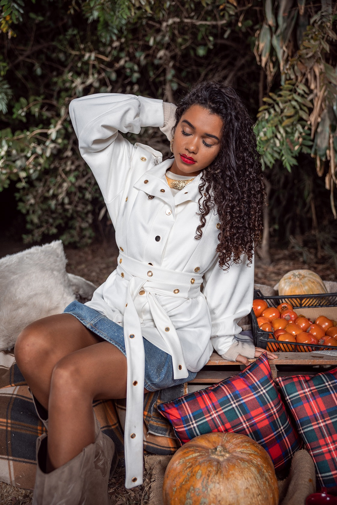 Woman in a white coat sitting outdoors with pumpkins and a basket of tomatoes.