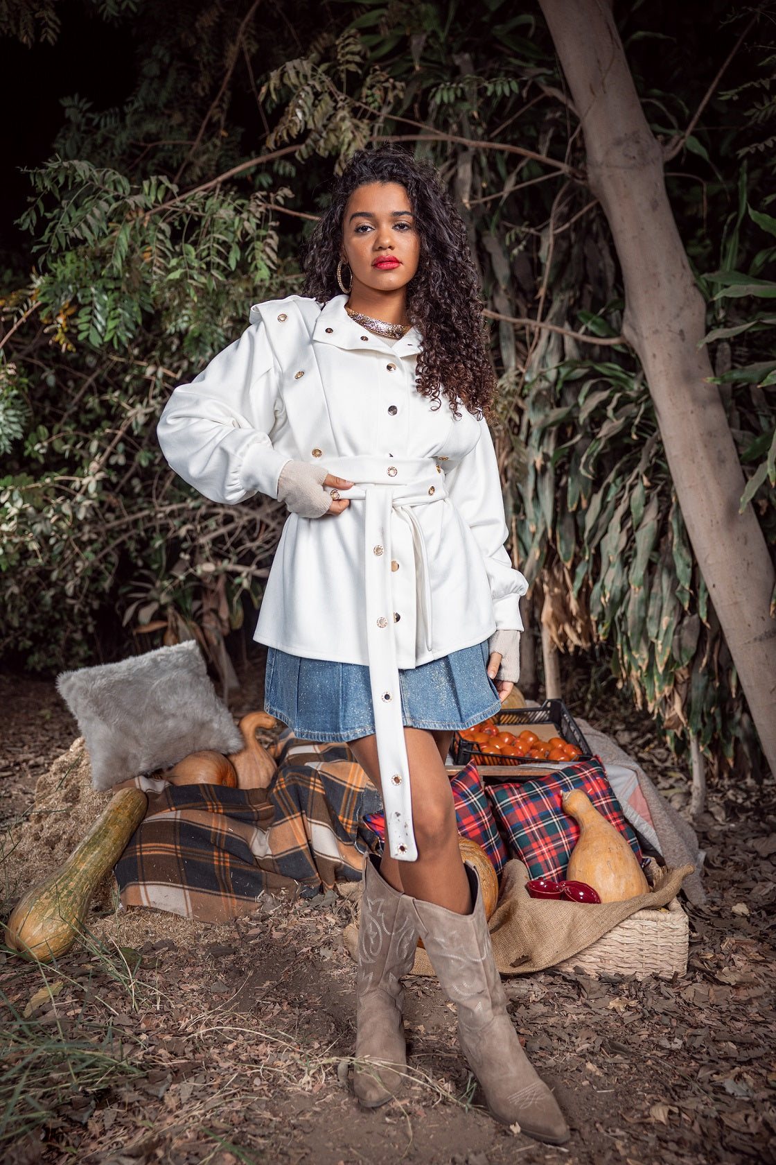 Woman in a white coat and skirt standing outdoors with a plaid blanket and pumpkins.