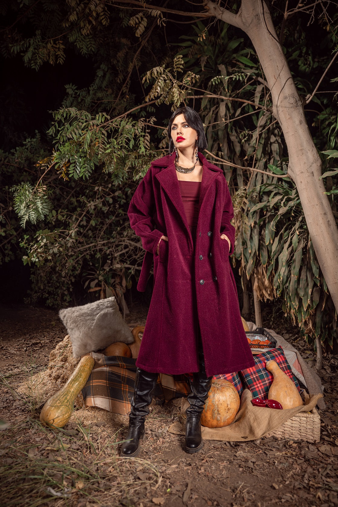 Woman in a long burgundy coat standing outdoors with pumpkins and autumn decorations.