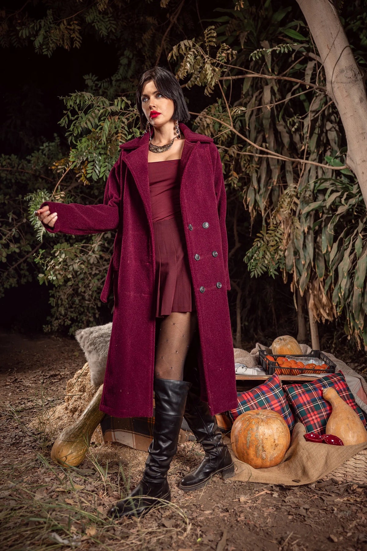 Woman in a long purple coat standing in a forest with pumpkins and a plaid blanket.