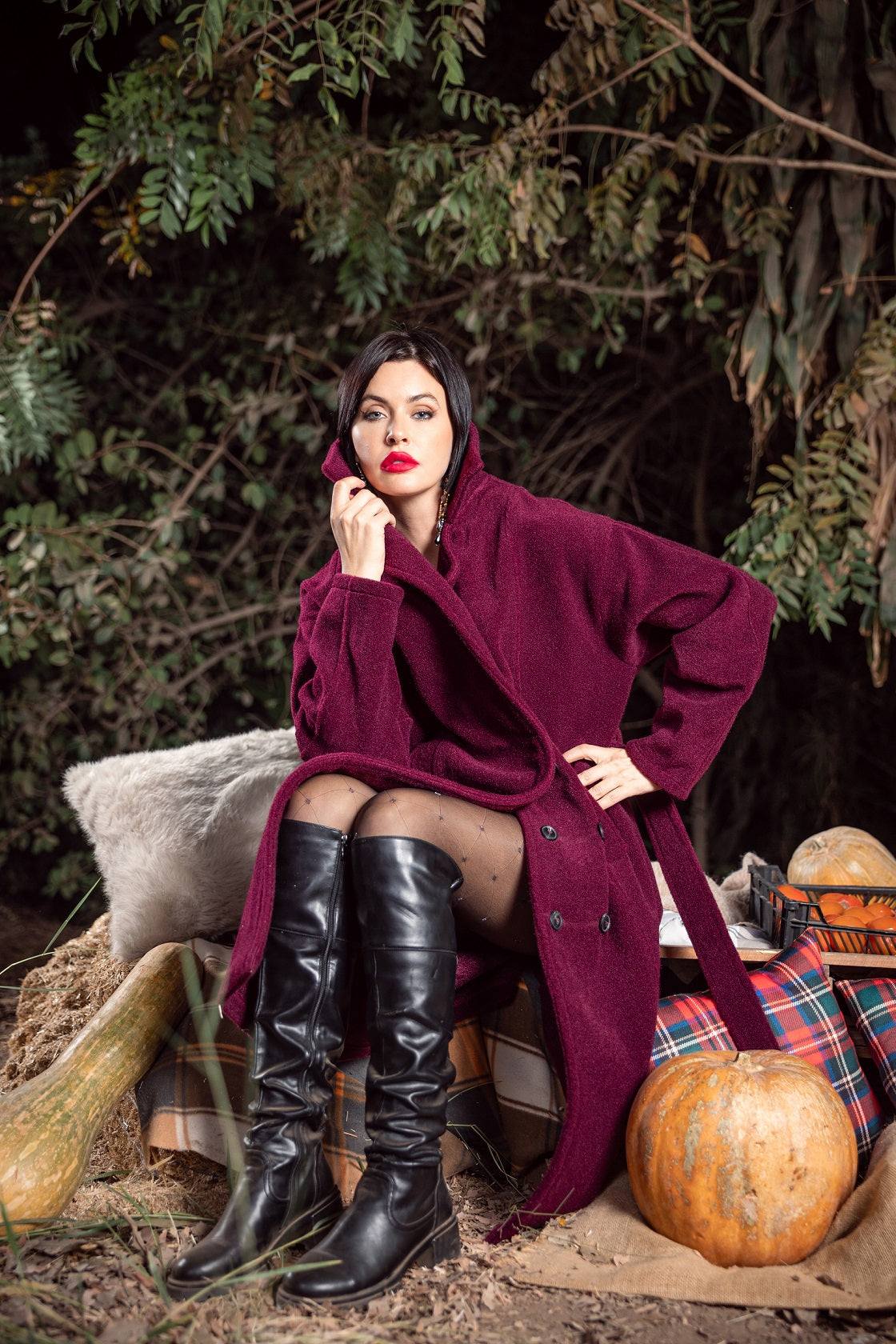 Woman in a purple coat and black knee-high boots sitting among pumpkins and autumn decorations.