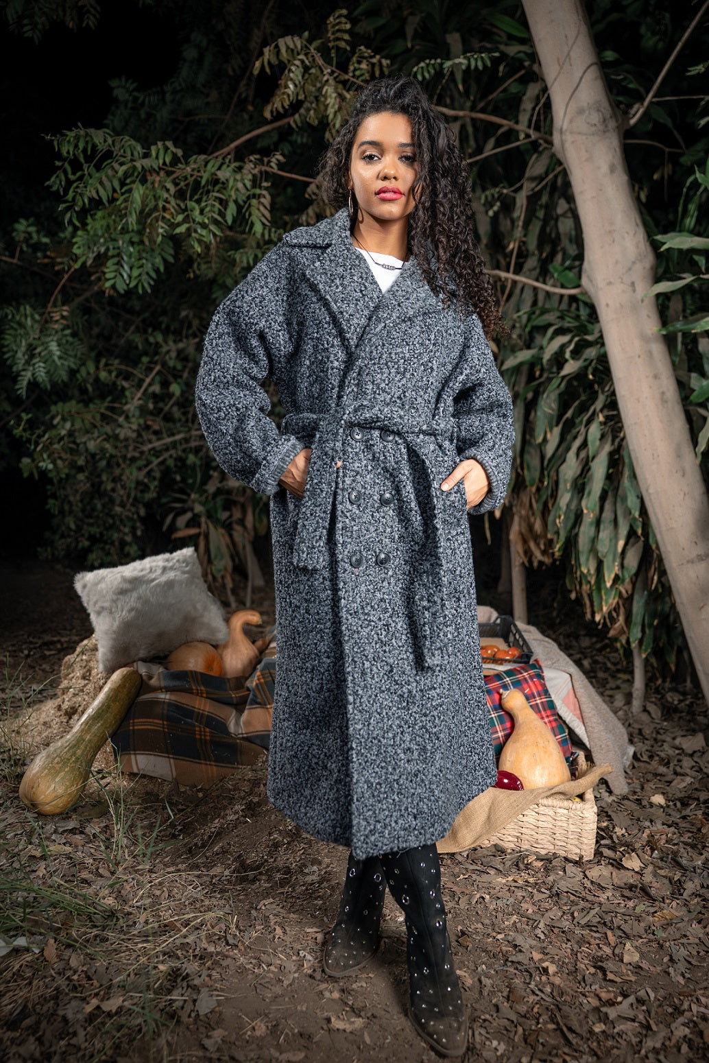 Woman wearing a long gray coat standing in a forest setting with baskets and pumpkins.