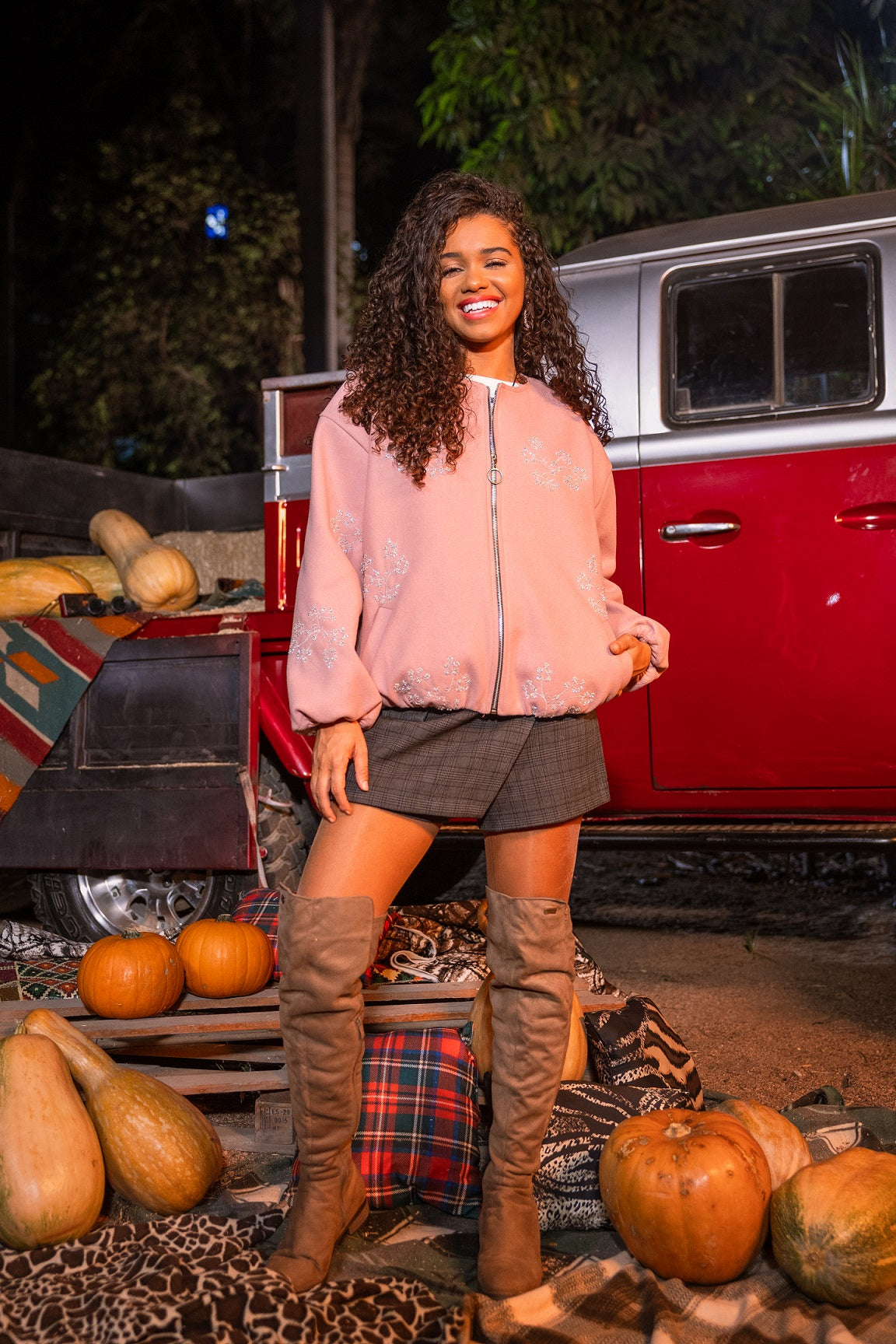 Woman standing in front of a red truck with pumpkins around, wearing a pink jacket and brown boots.