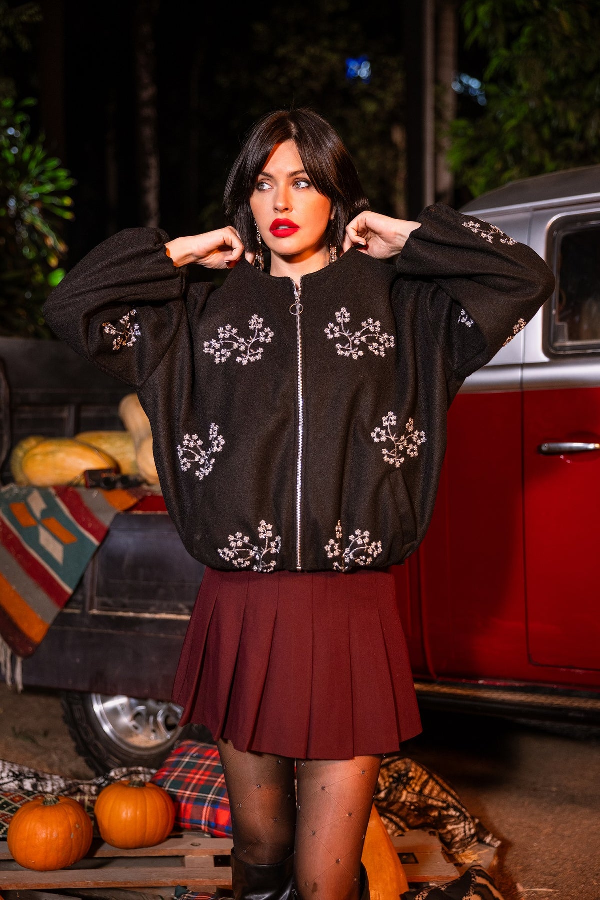 Woman wearing a black embroidered jacket in front of a vintage car at night.