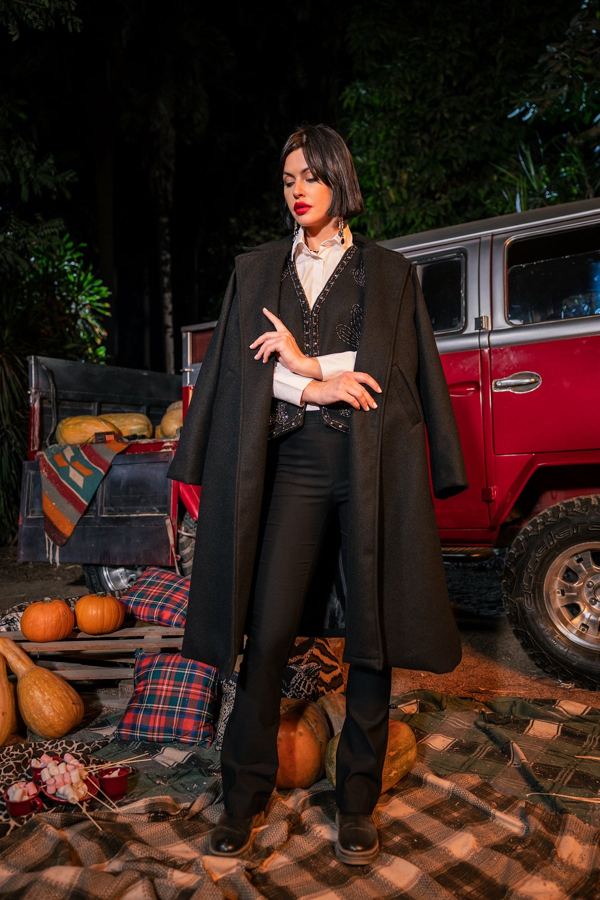 Woman in a long black coat standing in front of a red truck with pumpkins and autumn decorations.