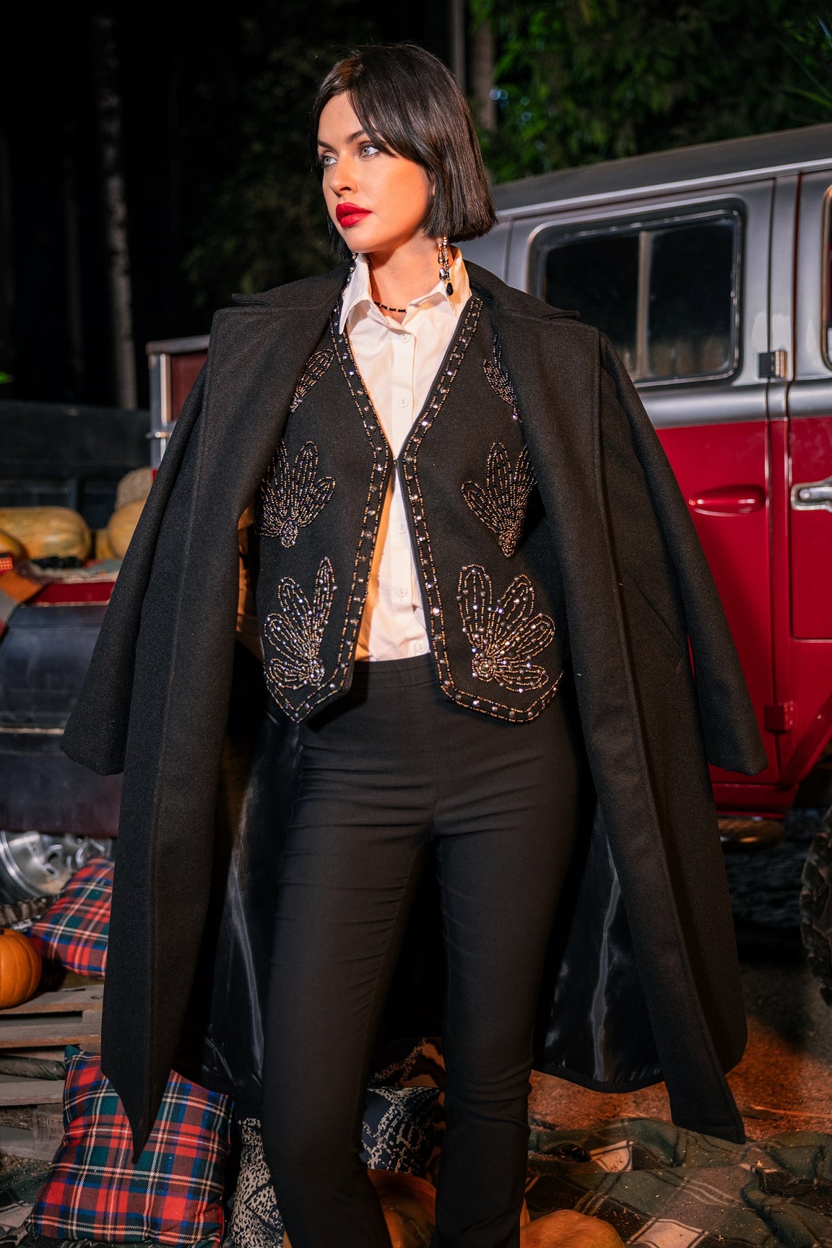 Woman in a long black coat with decorative elements standing in front of a vintage red vehicle.