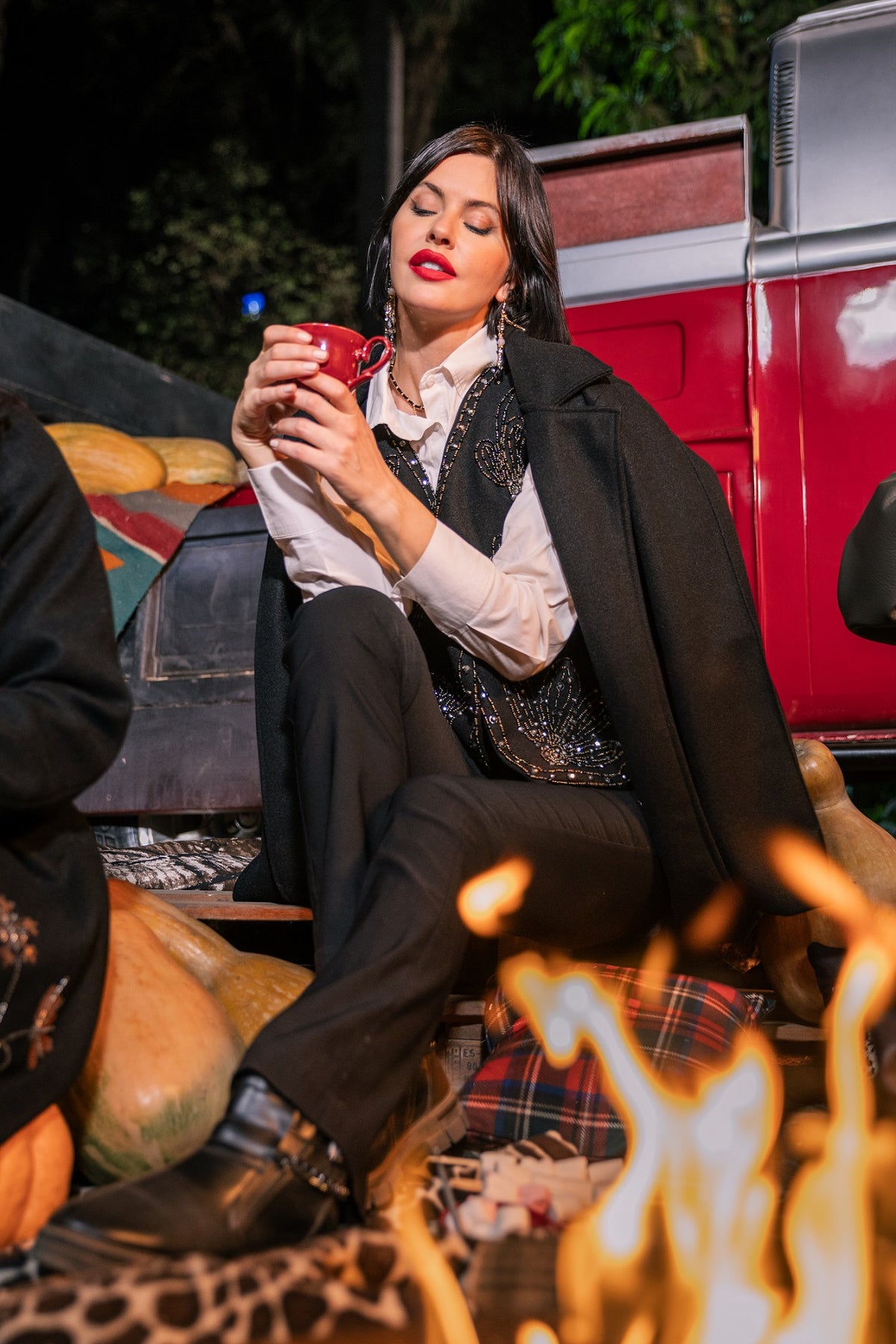 Woman sitting by a fire, holding a mug with a red truck in the background