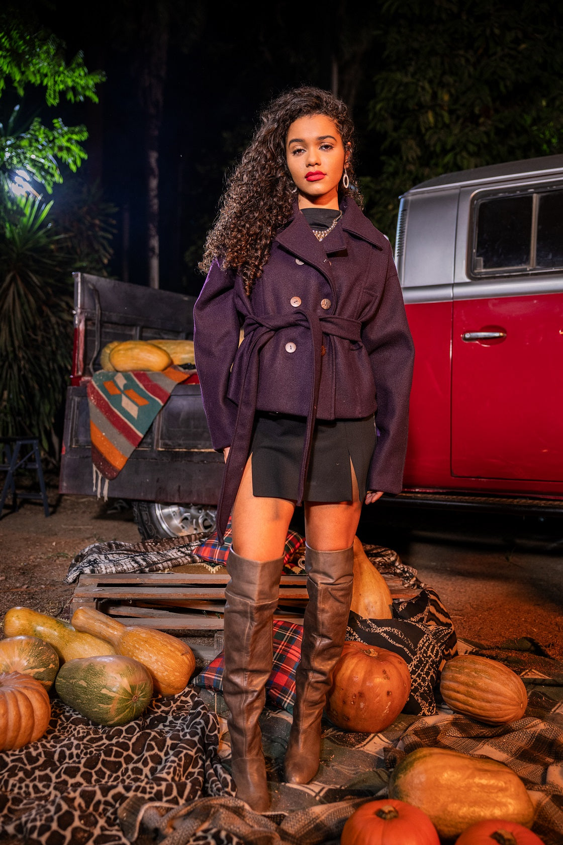 Woman in a purple coat standing in front of a red vehicle with pumpkins and gourds around.