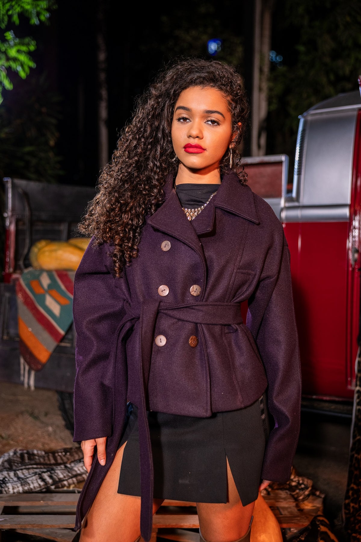Woman wearing a dark purple coat standing outdoors with a red vehicle and greenery in the background
