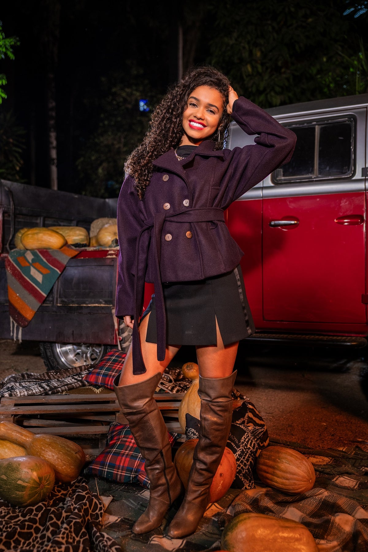 Woman in a dark coat and knee-high boots standing in front of a red vehicle with pumpkins around.