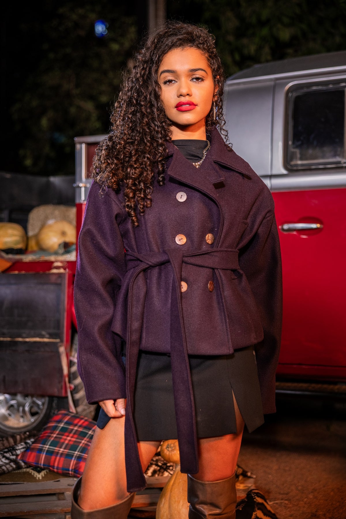 Woman in a dark coat standing in front of a vintage red vehicle at night.