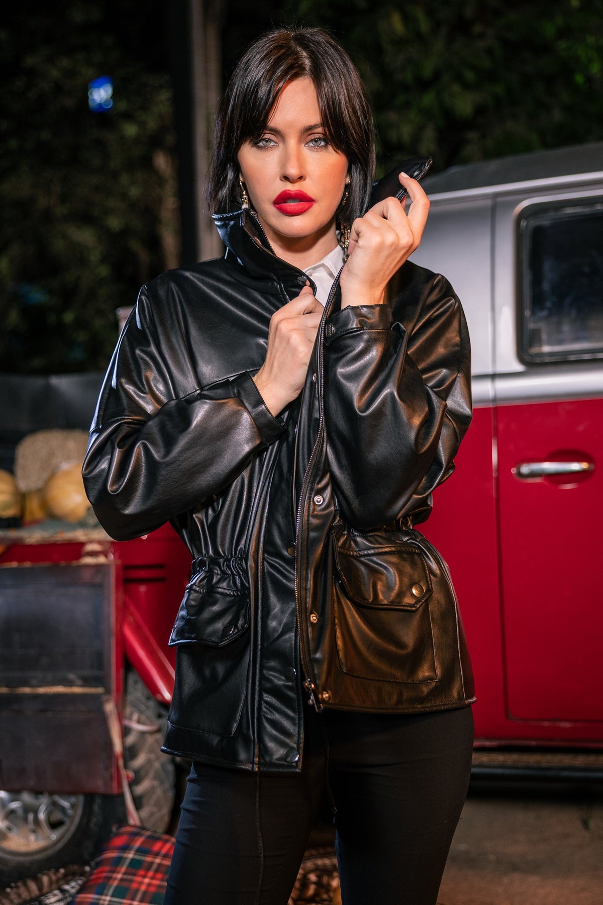 Woman in a black leather jacket standing in front of a red vehicle at night.