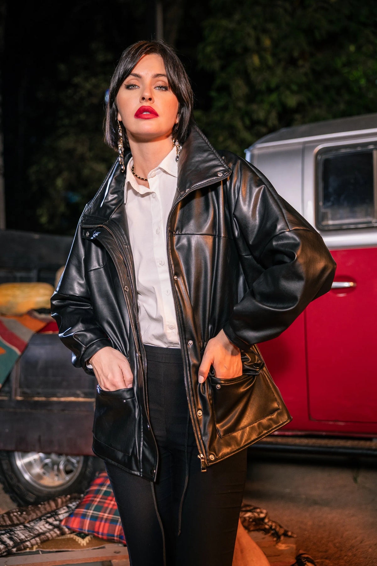 Woman in a black leather jacket standing in front of vintage cars at night.