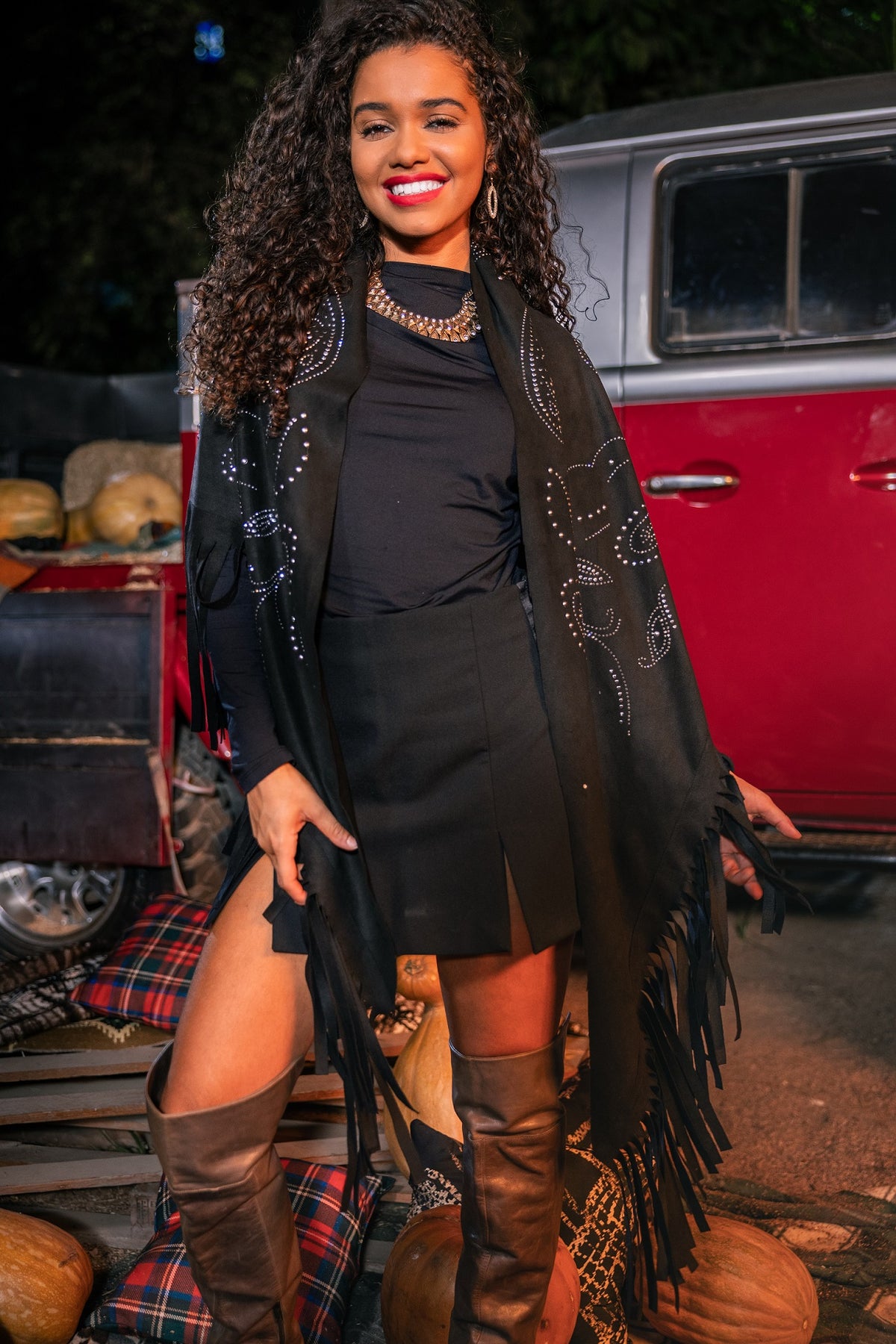 Woman in a black outfit with a fringe jacket standing in front of a vintage car.