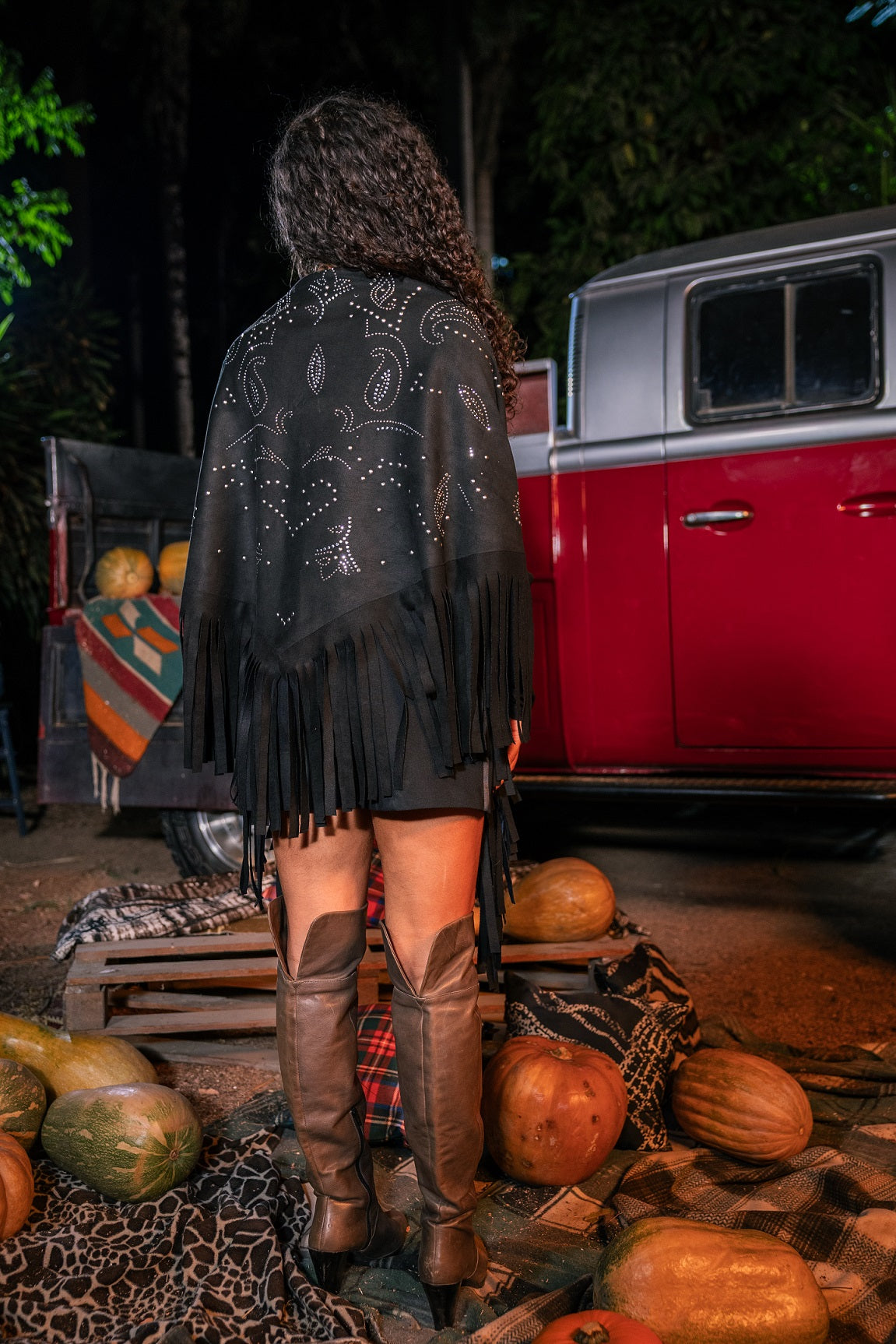 Person wearing a black fringe jacket and knee-high boots standing in front of a red truck with pumpkins around.