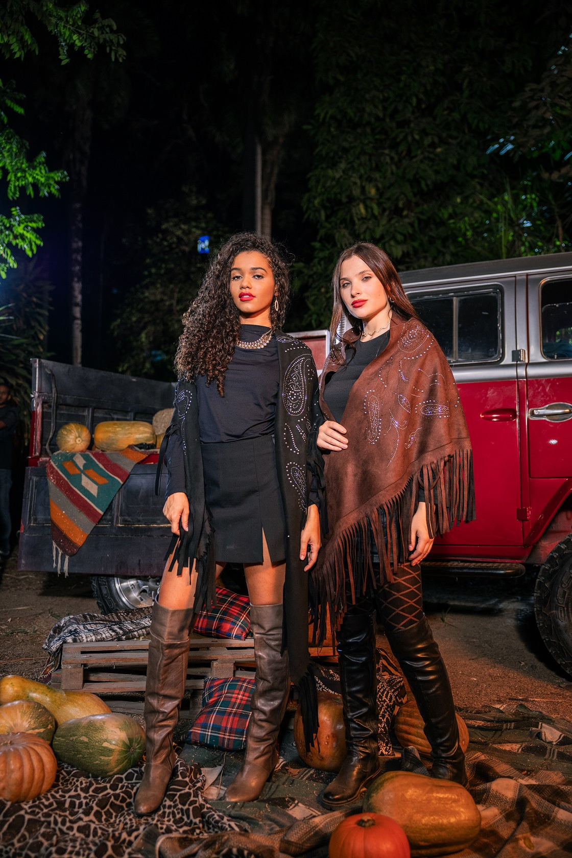 Two women standing in front of a red truck with pumpkins at night.