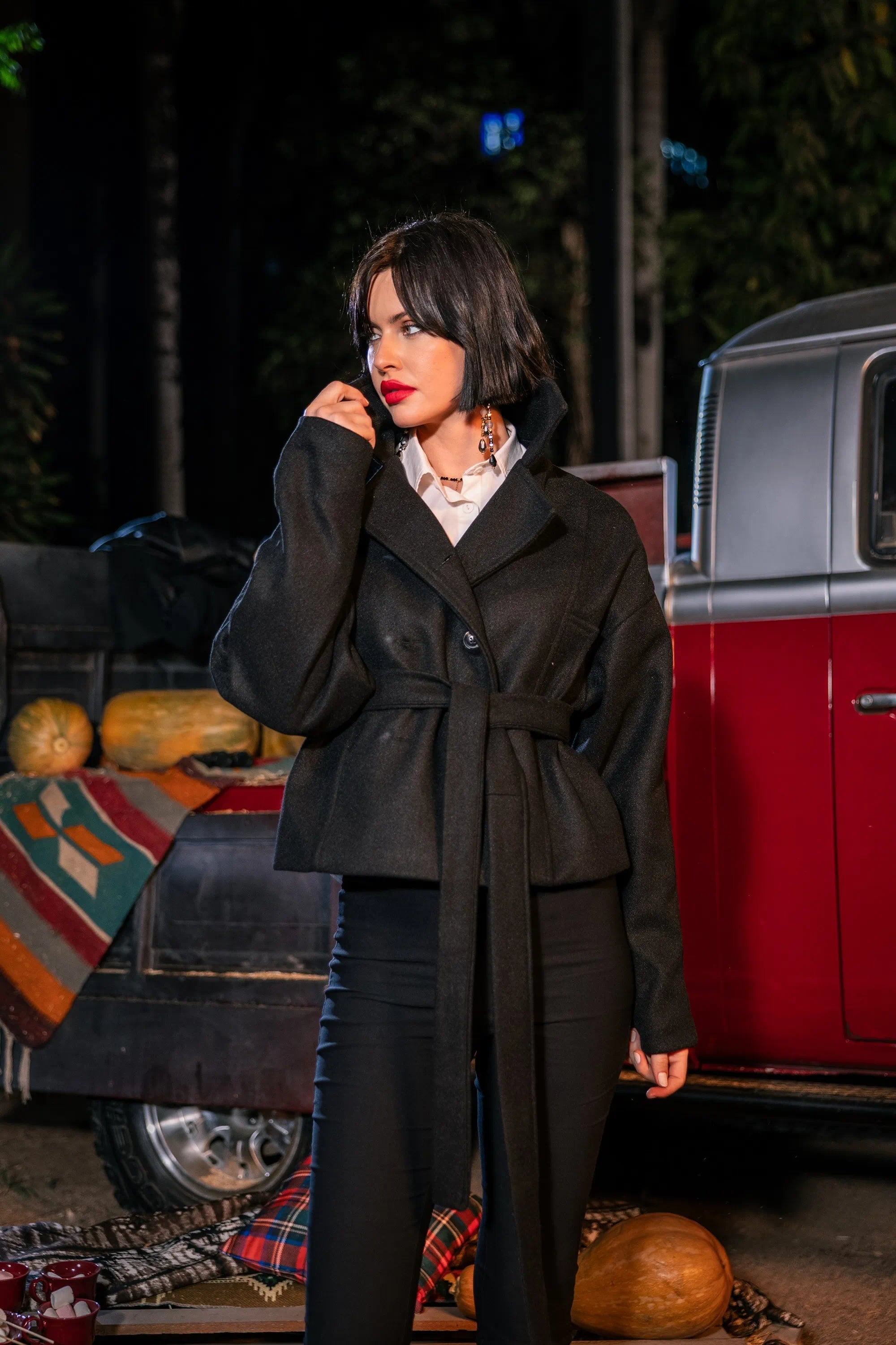 Woman in a long black coat standing in front of a red vehicle with autumn decorations.