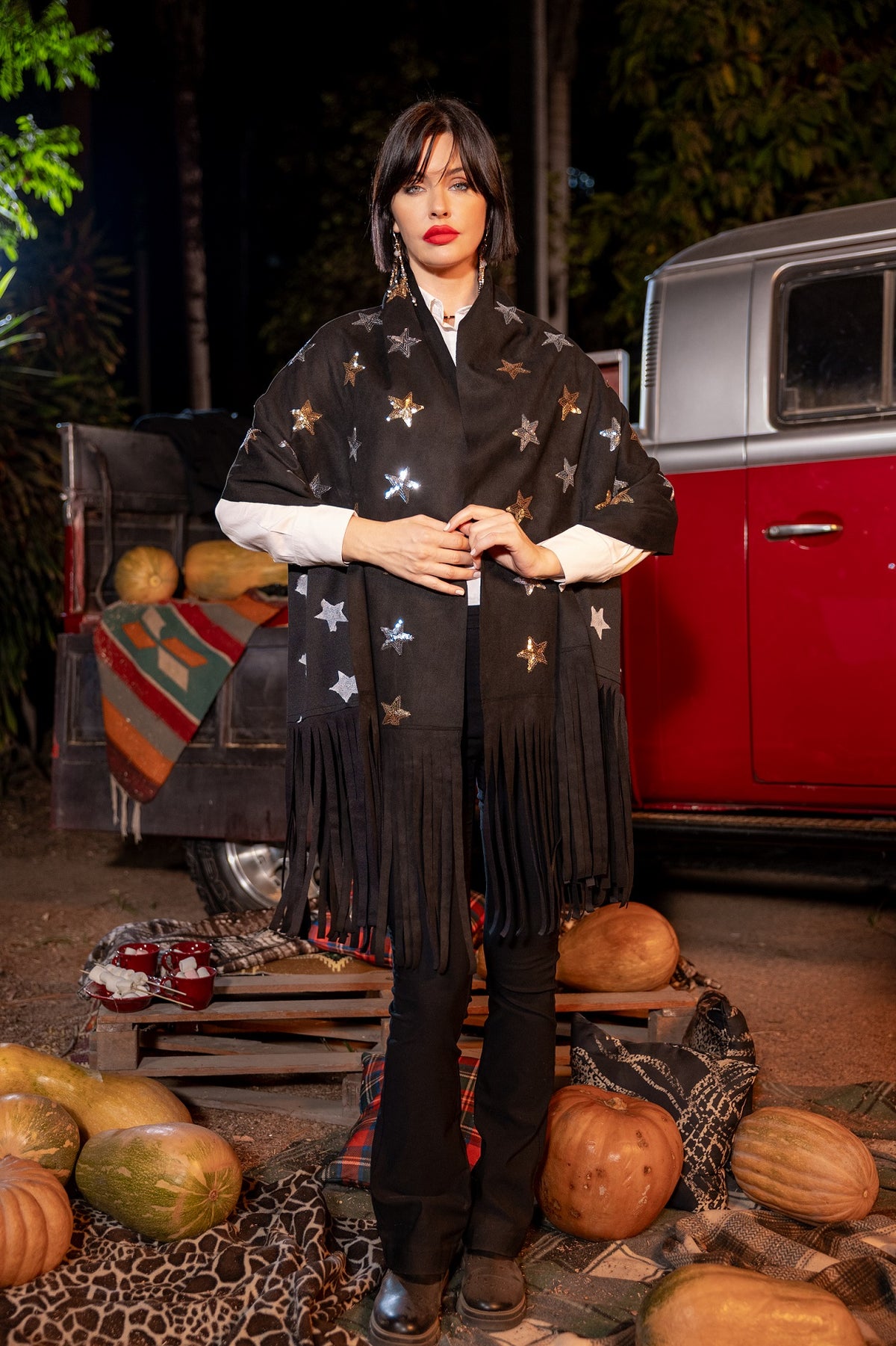 Woman wearing a black star-patterned poncho standing in front of a vintage car with pumpkins around.
