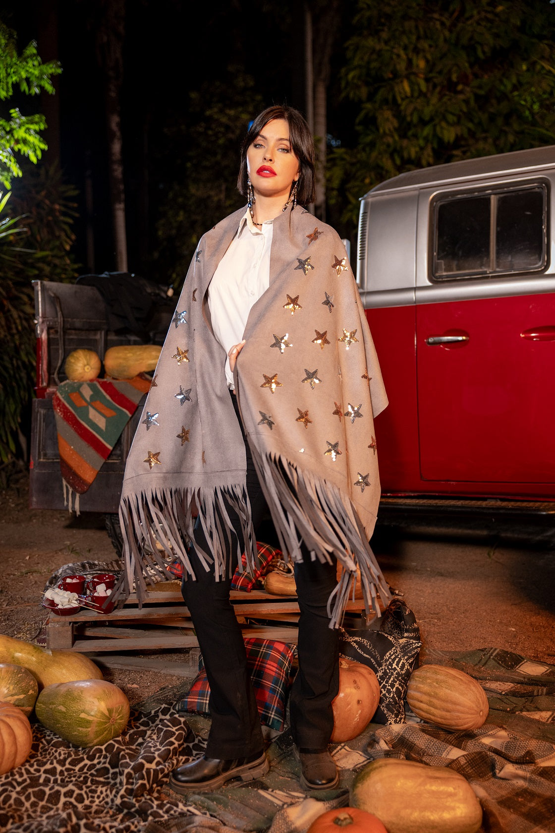 Woman wearing a star-patterned shawl in front of a vintage red car with pumpkins around.