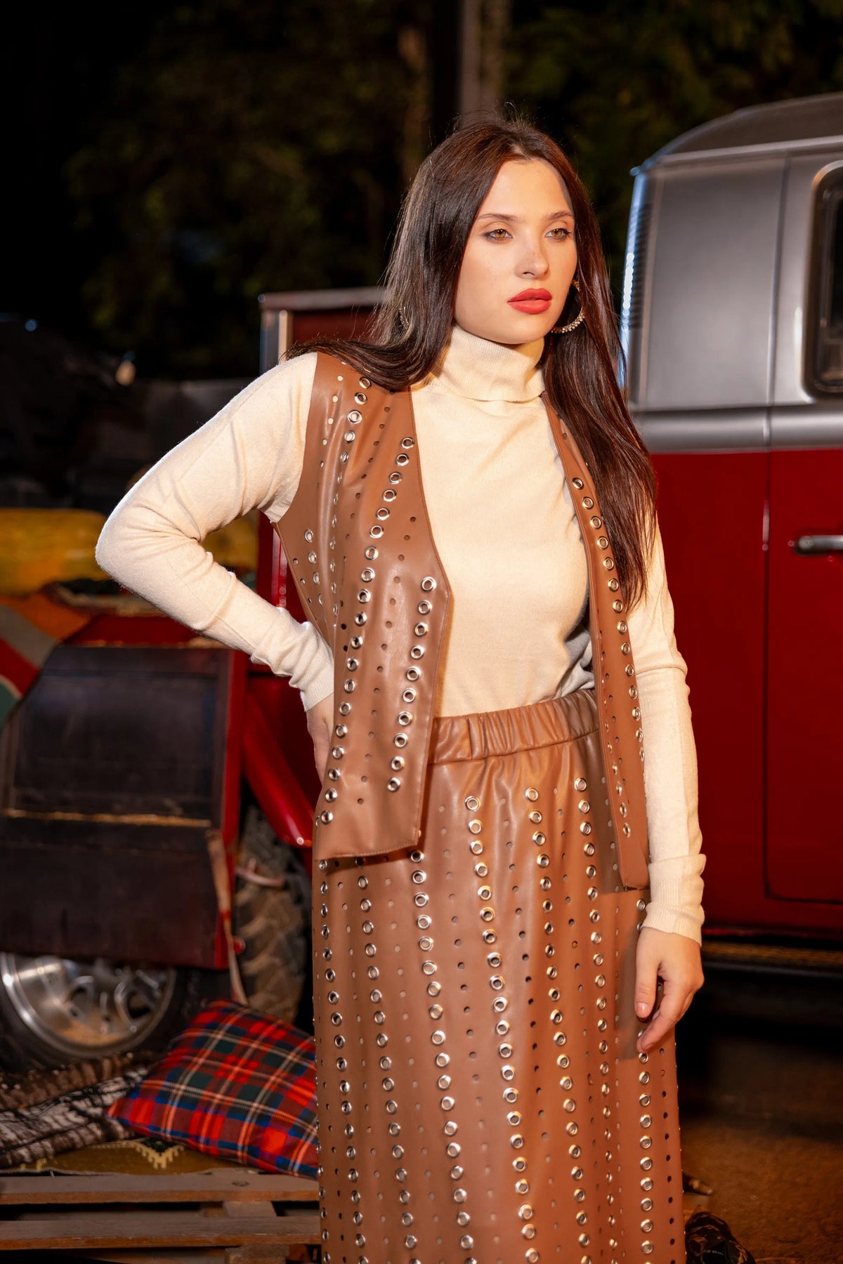 Woman wearing a brown studded vest and pants standing in front of a vintage red car.