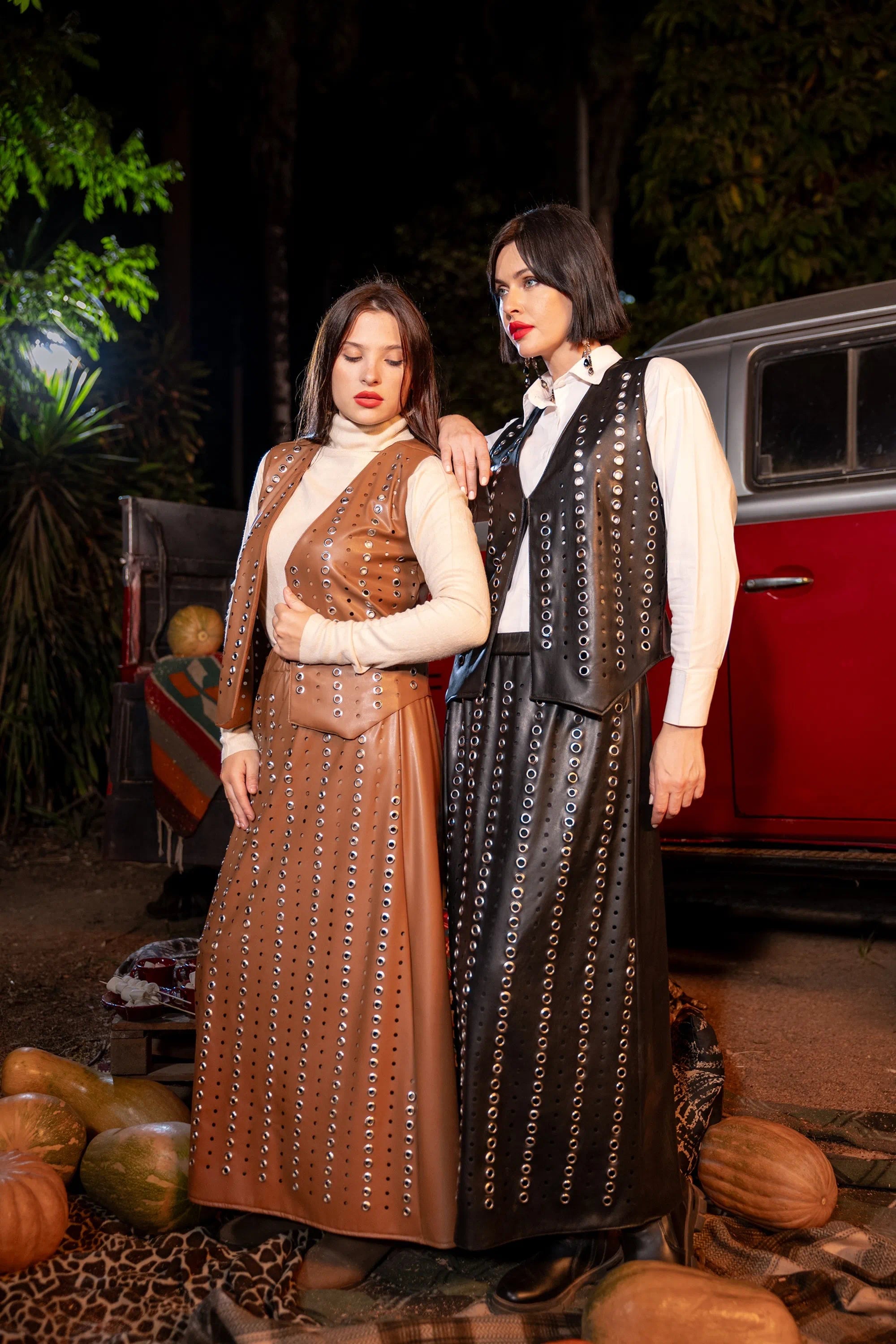 Two women in vintage-style outfits standing in front of a red car with pumpkins around.