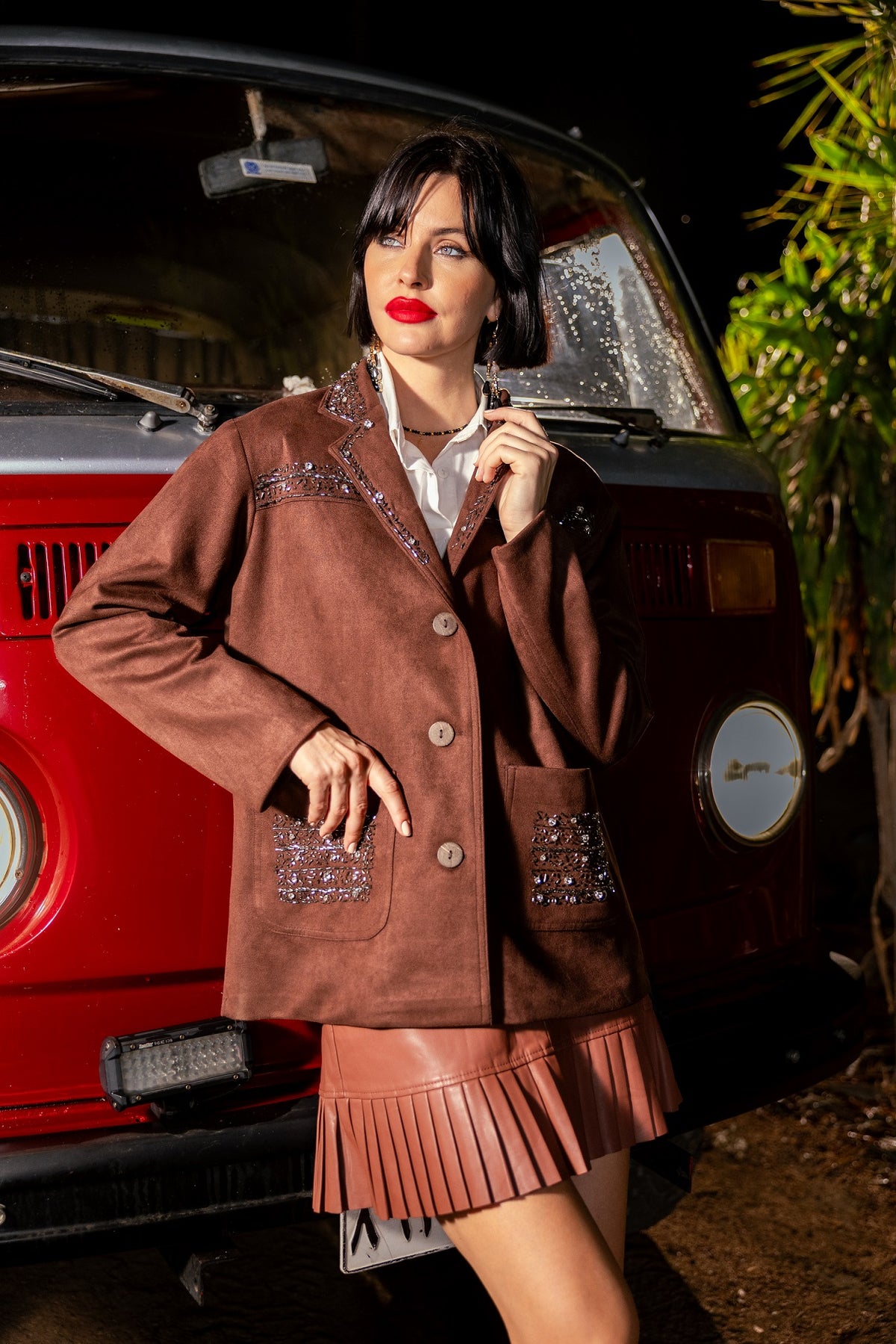 Woman in a brown jacket and skirt standing in front of a red car with a blurred background