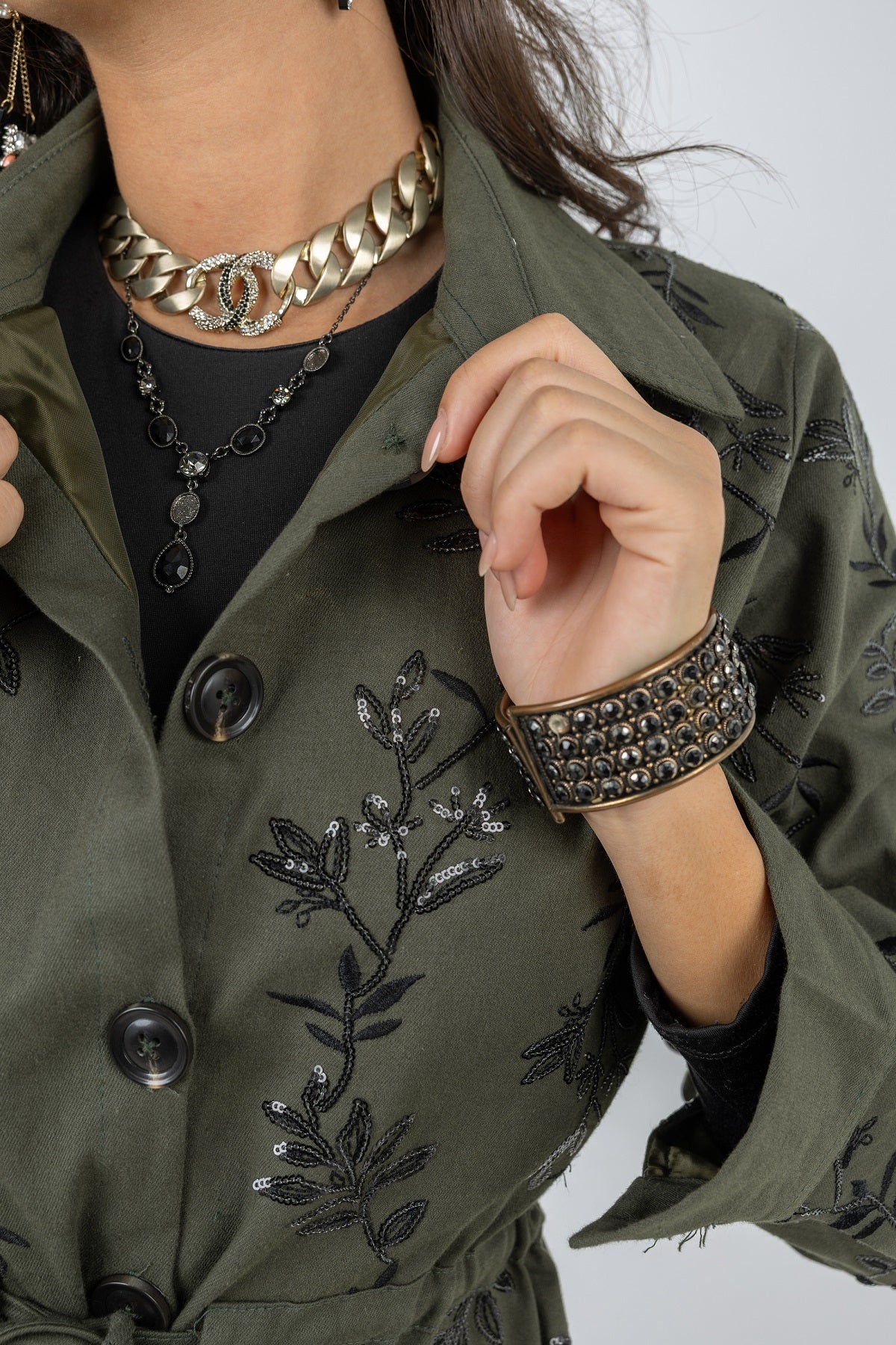 Person wearing a green embroidered jacket with floral patterns, black top, and gold chain necklace.